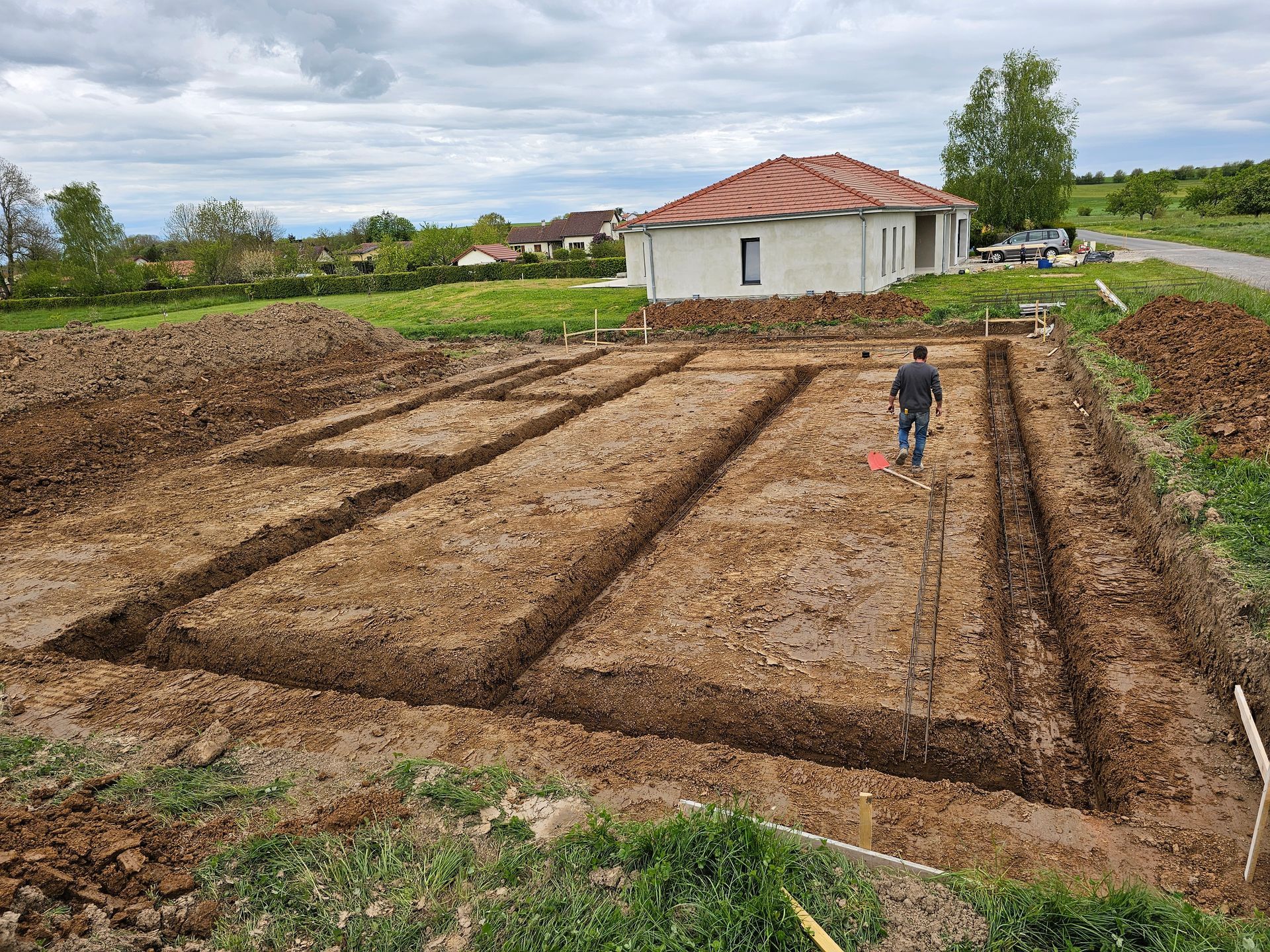 Des parterres de jardin fraîchement labourés, une personne marchant vers une maison blanche sous un ciel nuageux.