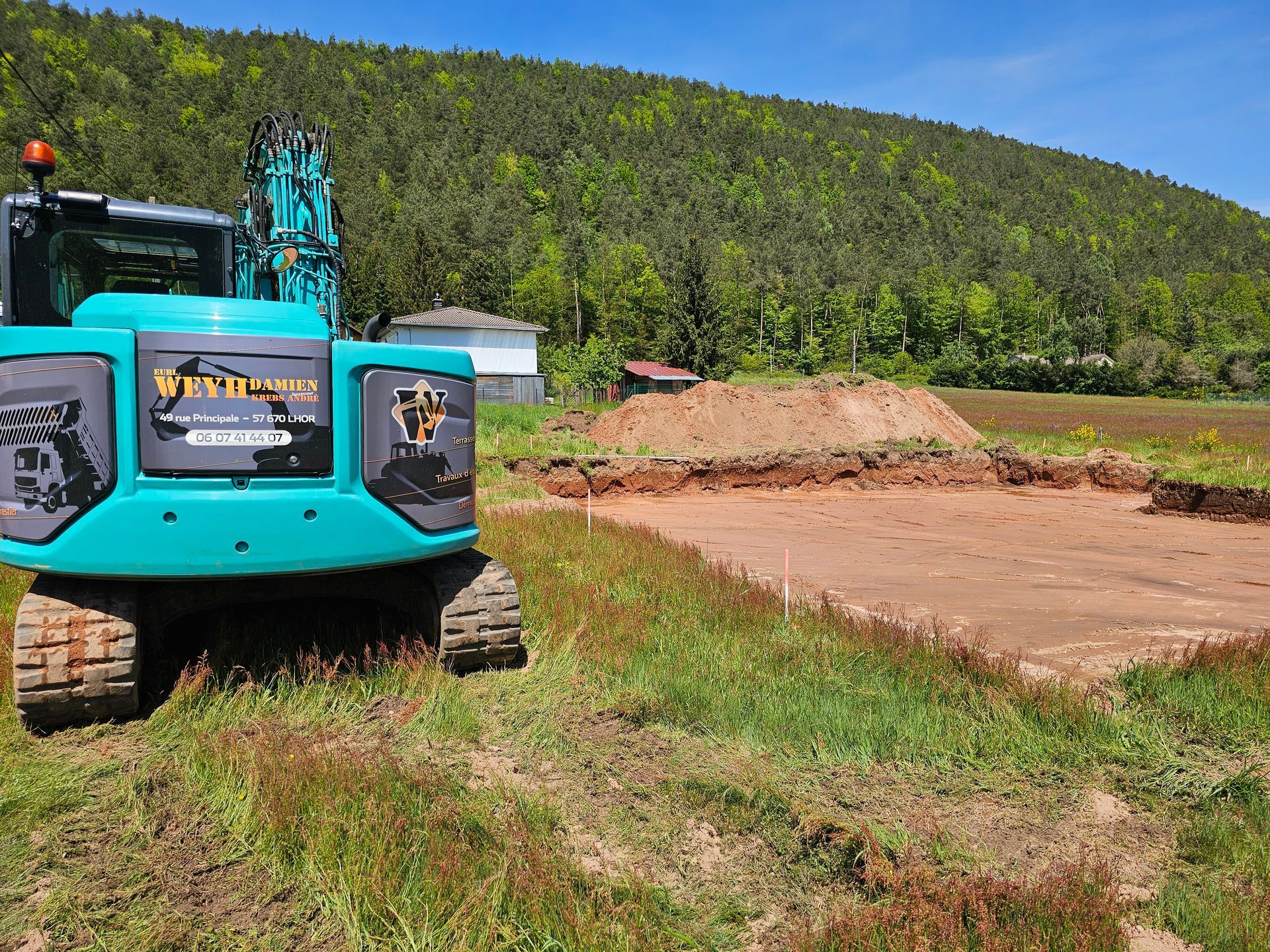 Une excavatrice turquoise est garée sur un terrain vague, avec une pelouse au premier plan et une colline boisée à l'arrière-plan.