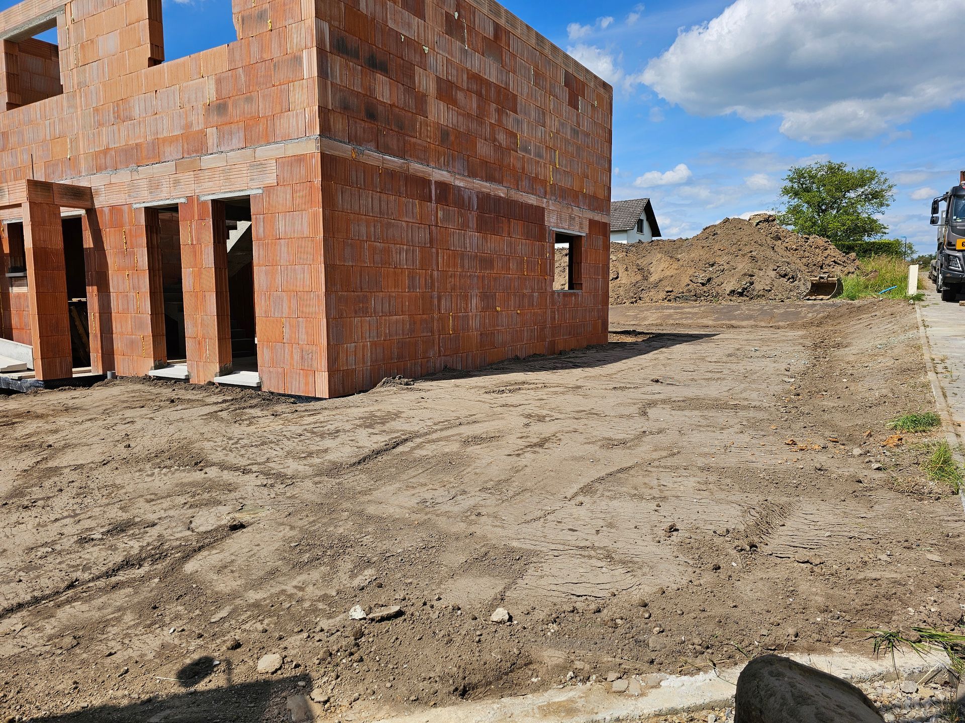 Bâtiment en briques en construction sur un terrain vague avec des tas de terre et un ciel partiellement nuageux.