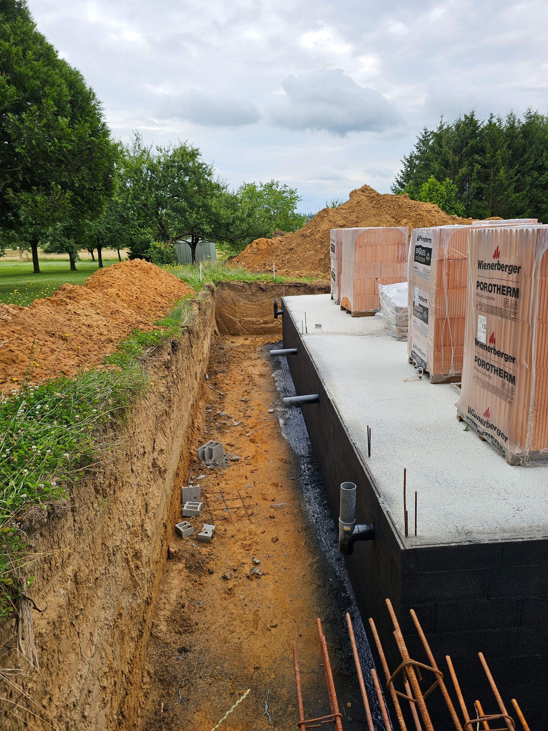 Chantier de construction avec murs de fondation, tranchée et tas de terre près des arbres.