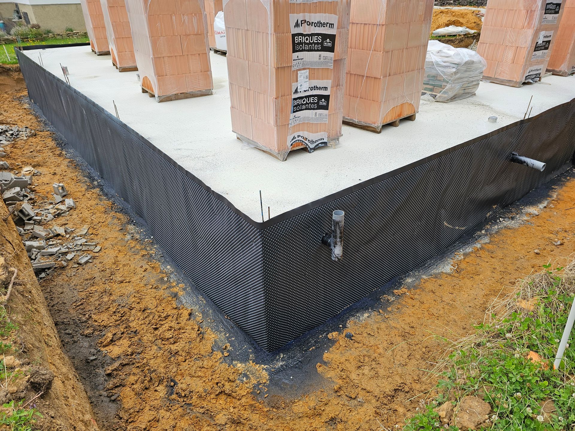 Fondations en béton noir étanche avec colonnes en briques sur un terrain en terre battue en construction.