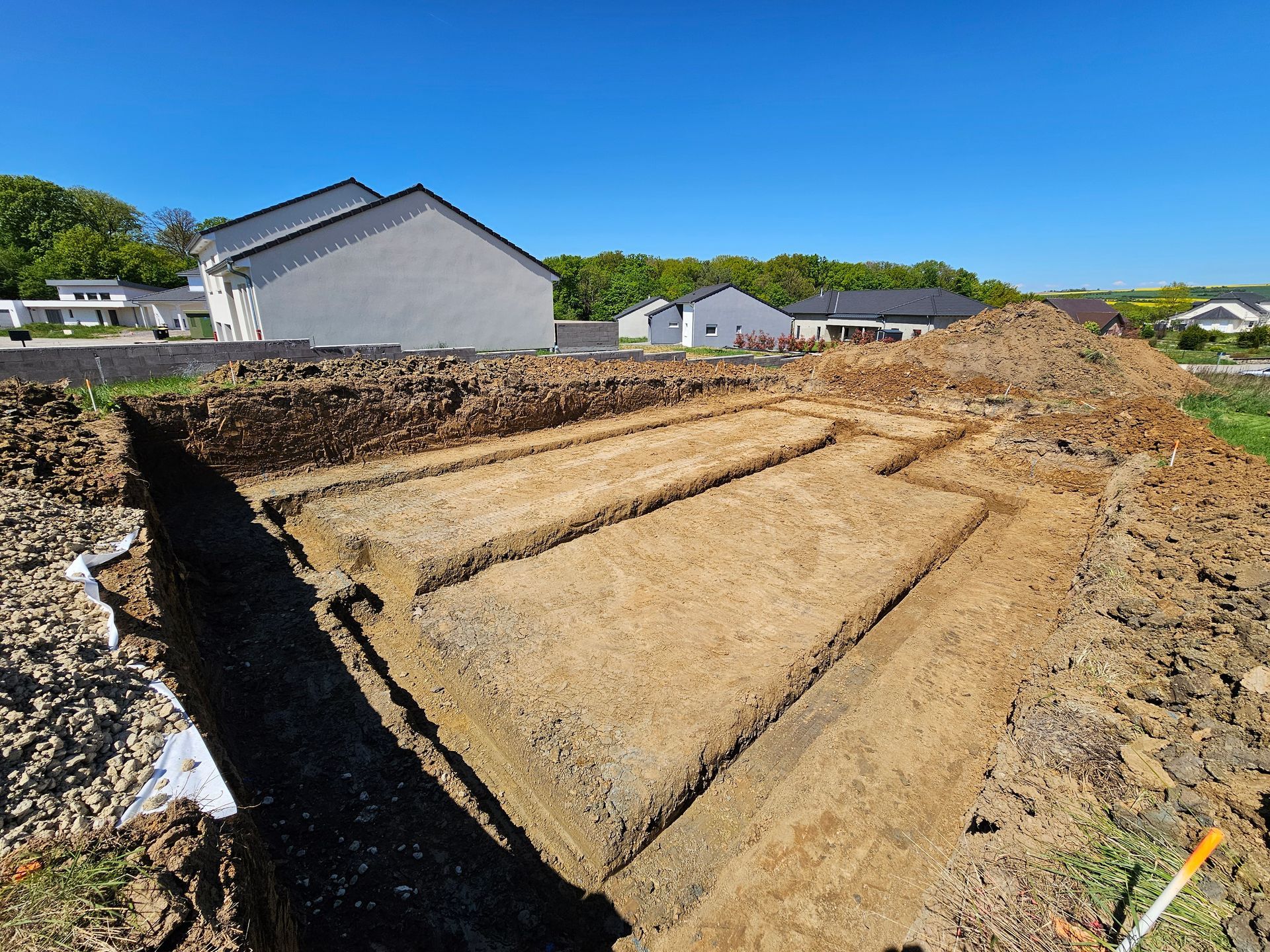Tranchée de fondation creusée sur un chantier de construction avec des maisons en arrière-plan sous un ciel dégagé.