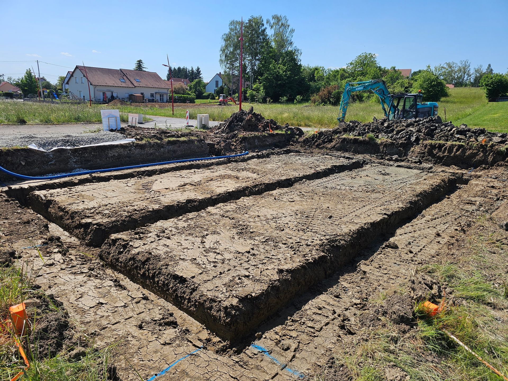 Une excavatrice nivelle un grand terrain en terre battue à côté d'un champ et de maisons sous un ciel dégagé.