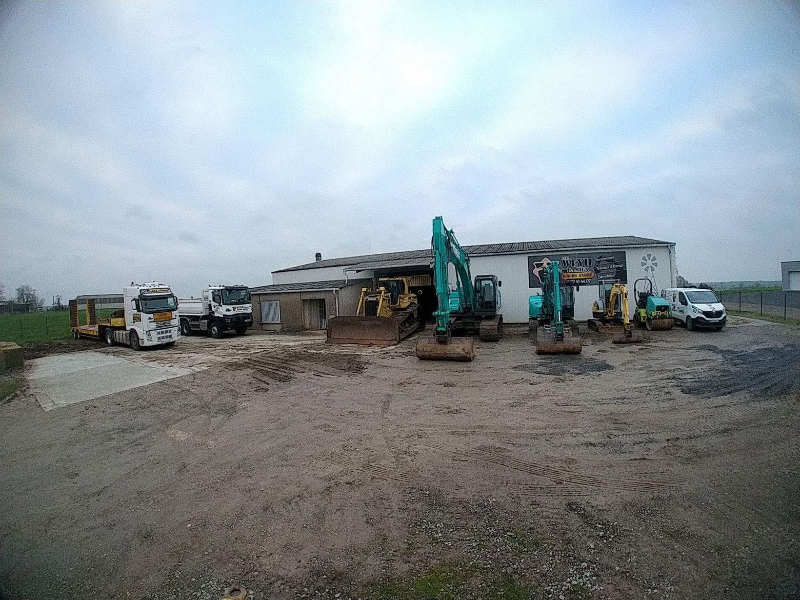 Une pelleteuse verte creuse une tranchée à côté d'une maison en briques sur un chantier boueux.