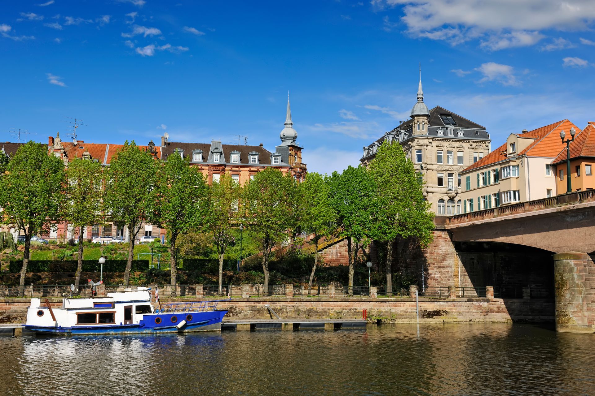 Ville riveraine avec des quais bordés d'arbres, des bateaux et un pont sous un ciel bleu.