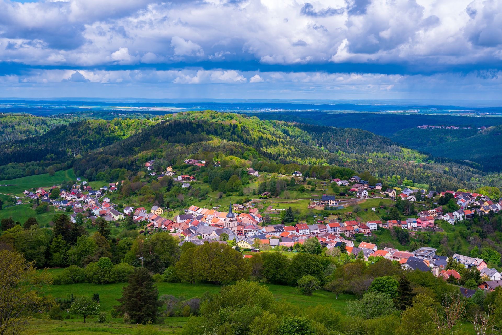 Village dans une vallée verdoyante aux maisons aux toits rouges, aux collines ondulantes et au ciel bleu nuageux.