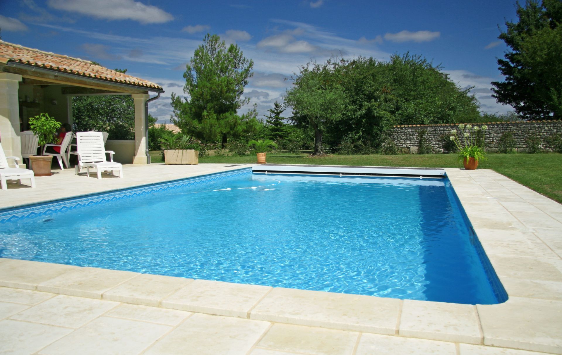 Piscine dans le jardin avec eau bleue, terrasse en pierre blanche et verdure sous un ciel ensoleillé.