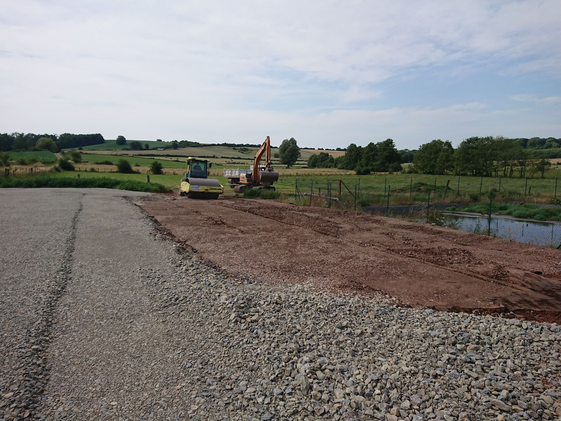 Travaux routiers avec une excavatrice et un camion à côté d'un chemin de gravier près d'un étang dans un champ rural.