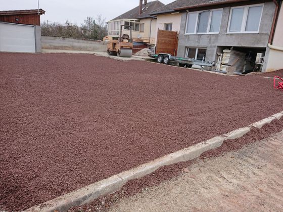 Terrain de construction recouvert de gravier, jouxtant des maisons, avec une pelleteuse stationnée par temps couvert.