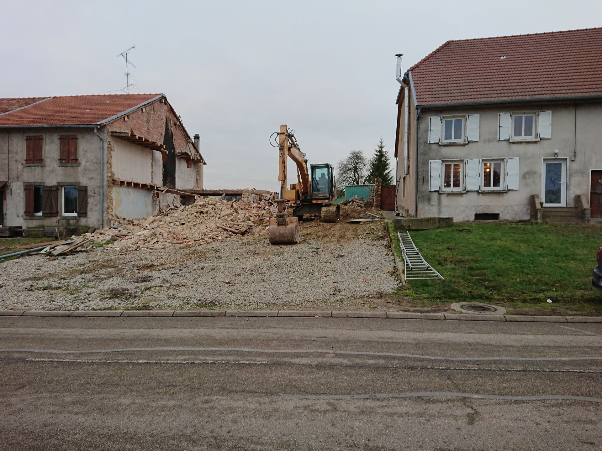 Une pelleteuse dégage les gravats entre deux maisons dans une rue résidentielle en construction.