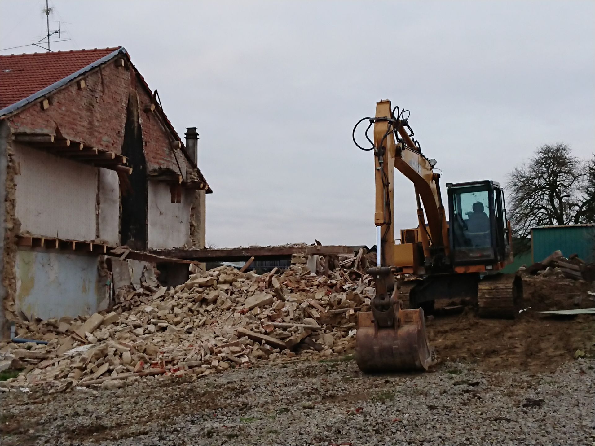 Une pelleteuse démolit une maison, les gravats s'entassent à côté du bâtiment en briques endommagé.