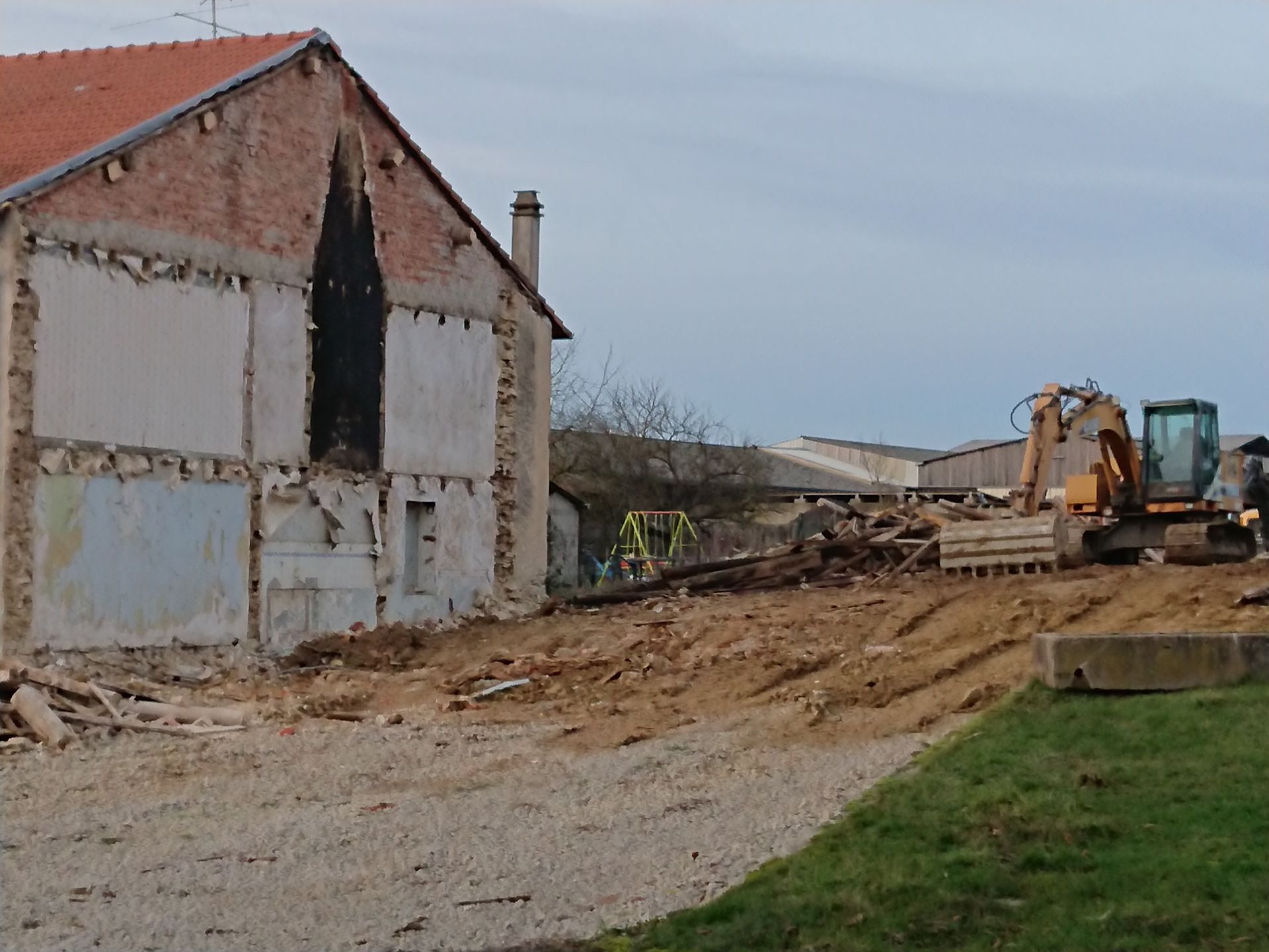 Site de démolition avec une maison partiellement démolie, des gravats et une excavatrice sur un terrain en terre battue.