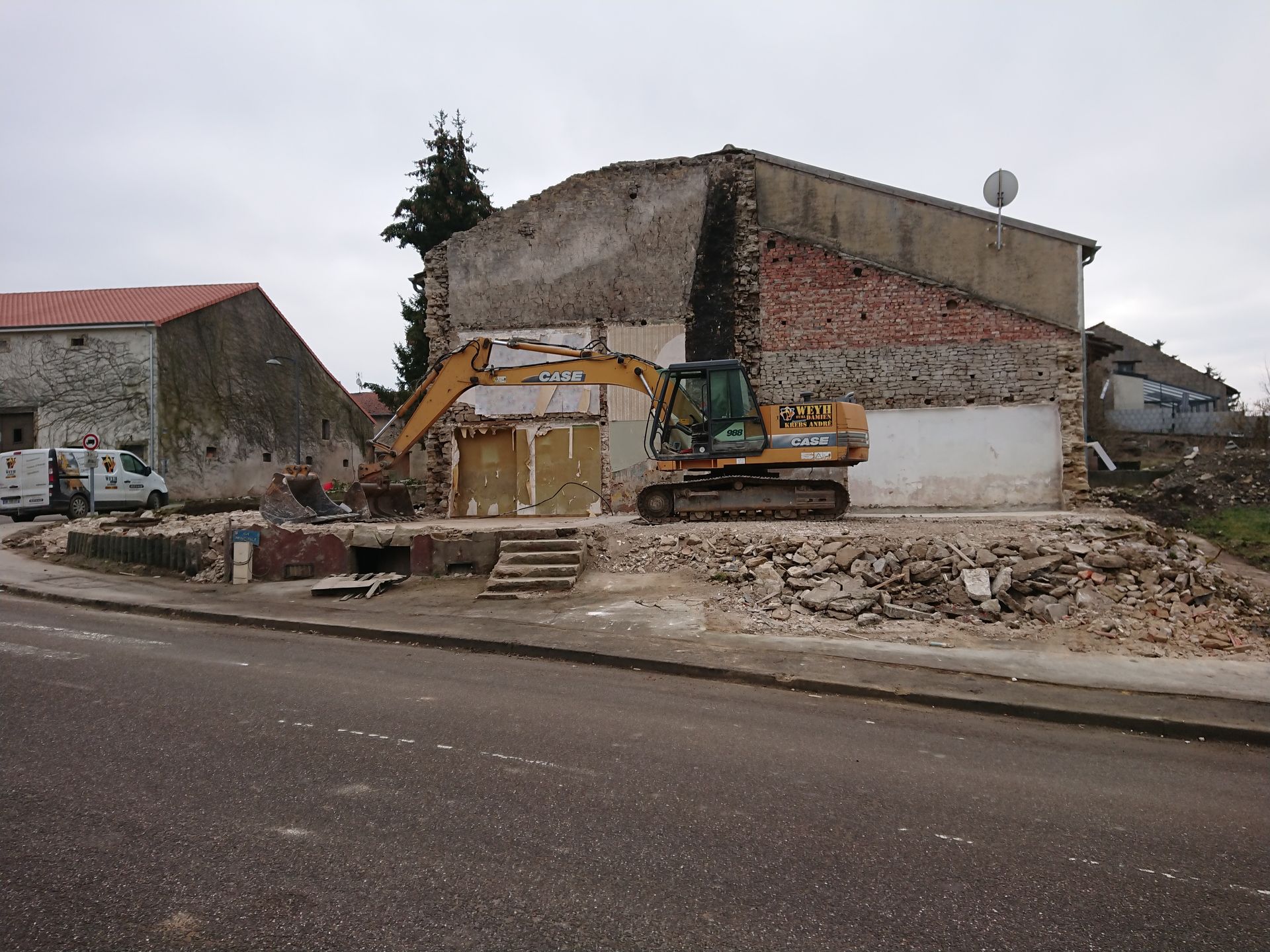 Une pelleteuse démolit un bâtiment en briques en bordure de route, des gravats s'accumulant devant.