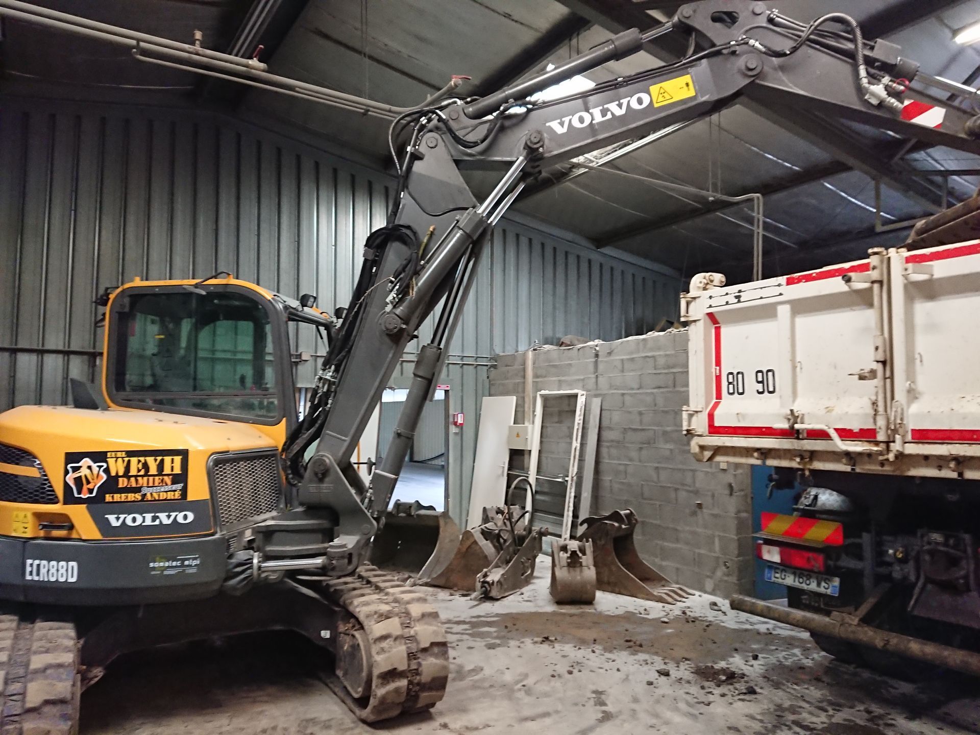 Une pelle mécanique jaune se trouve dans une station de lavage pour camions, à côté d'un camion-benne blanc sous une flèche levée.