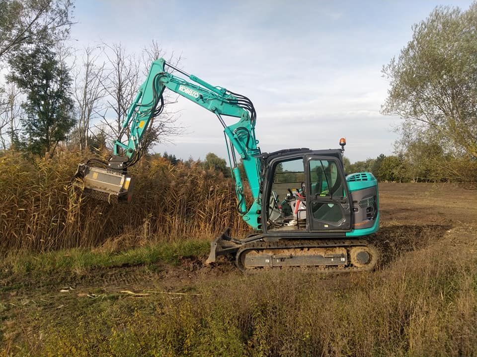 Une pelleteuse turquoise débroussaillant un champ boueux sous un ciel nuageux.