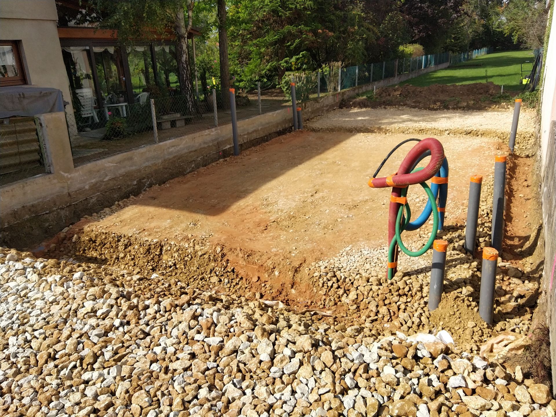 Chantier de construction extérieur avec zone de terre excavée, gravier et canalisations apparentes avec tuyaux colorés.
