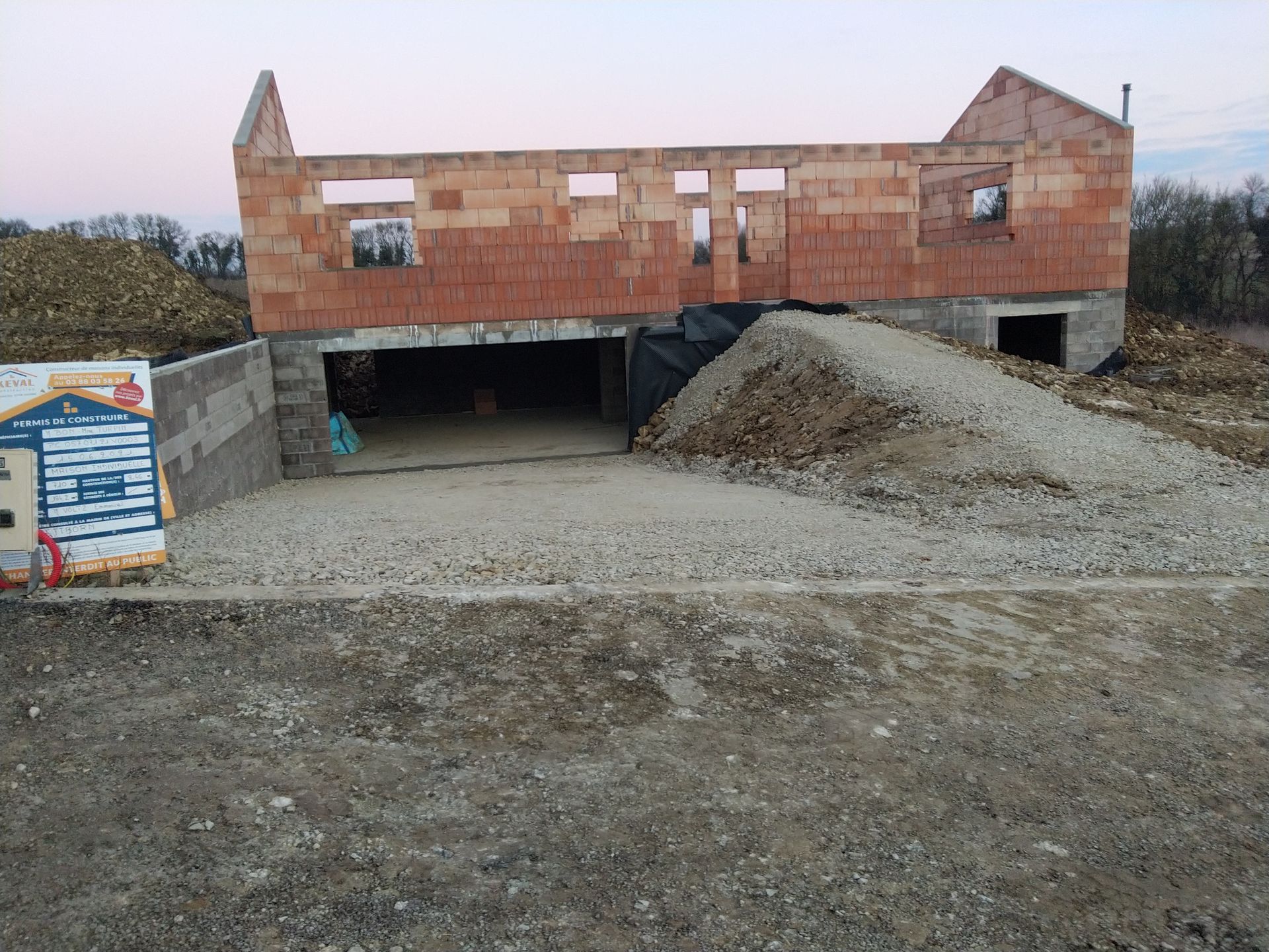 Maison en briques partiellement construite avec des tas de gravier et une entrée inachevée sur un terrain en terre battue au crépuscule.