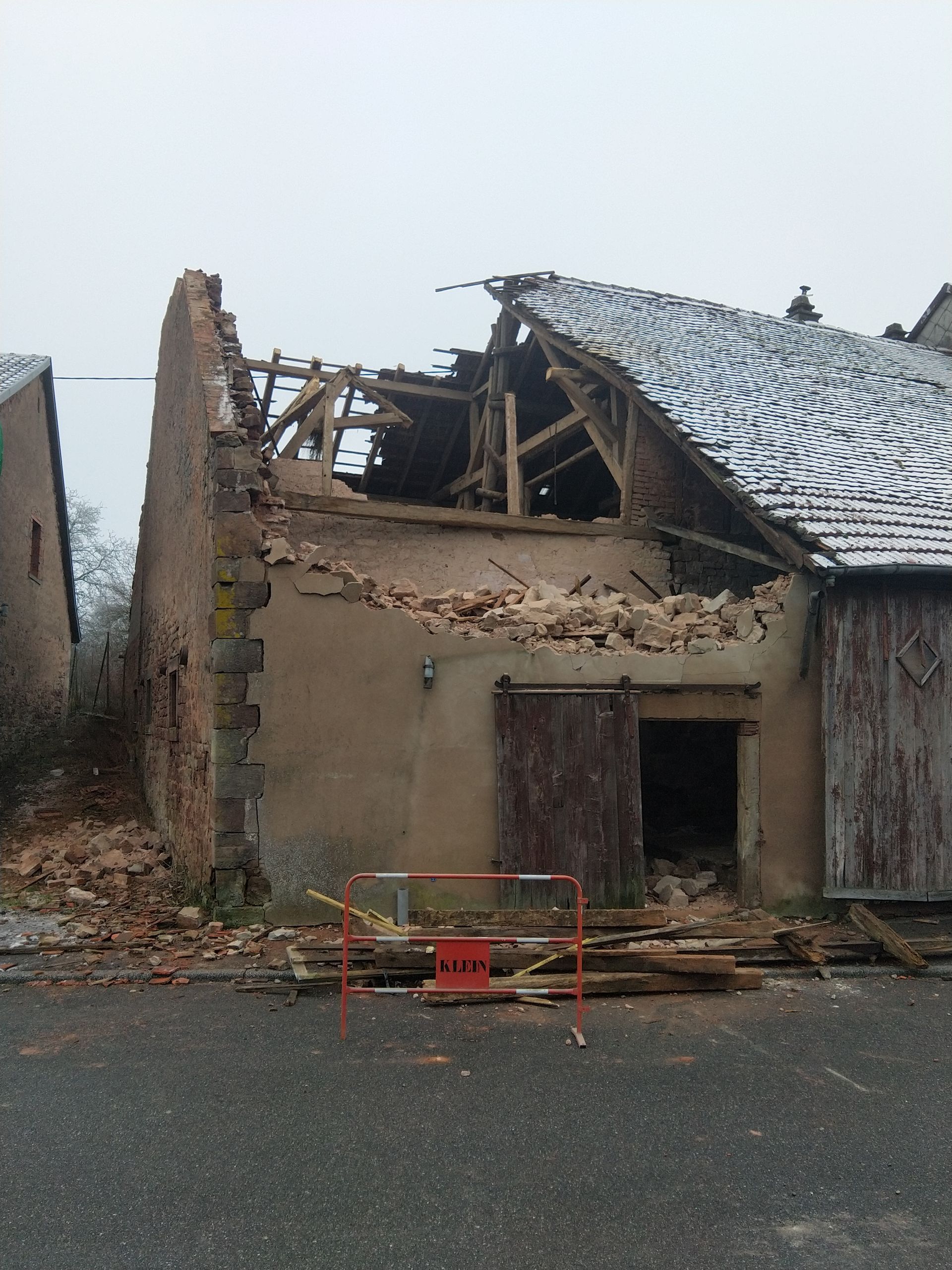 Maison effondrée, toiture gravement endommagée et débris devant, rue mouillée.