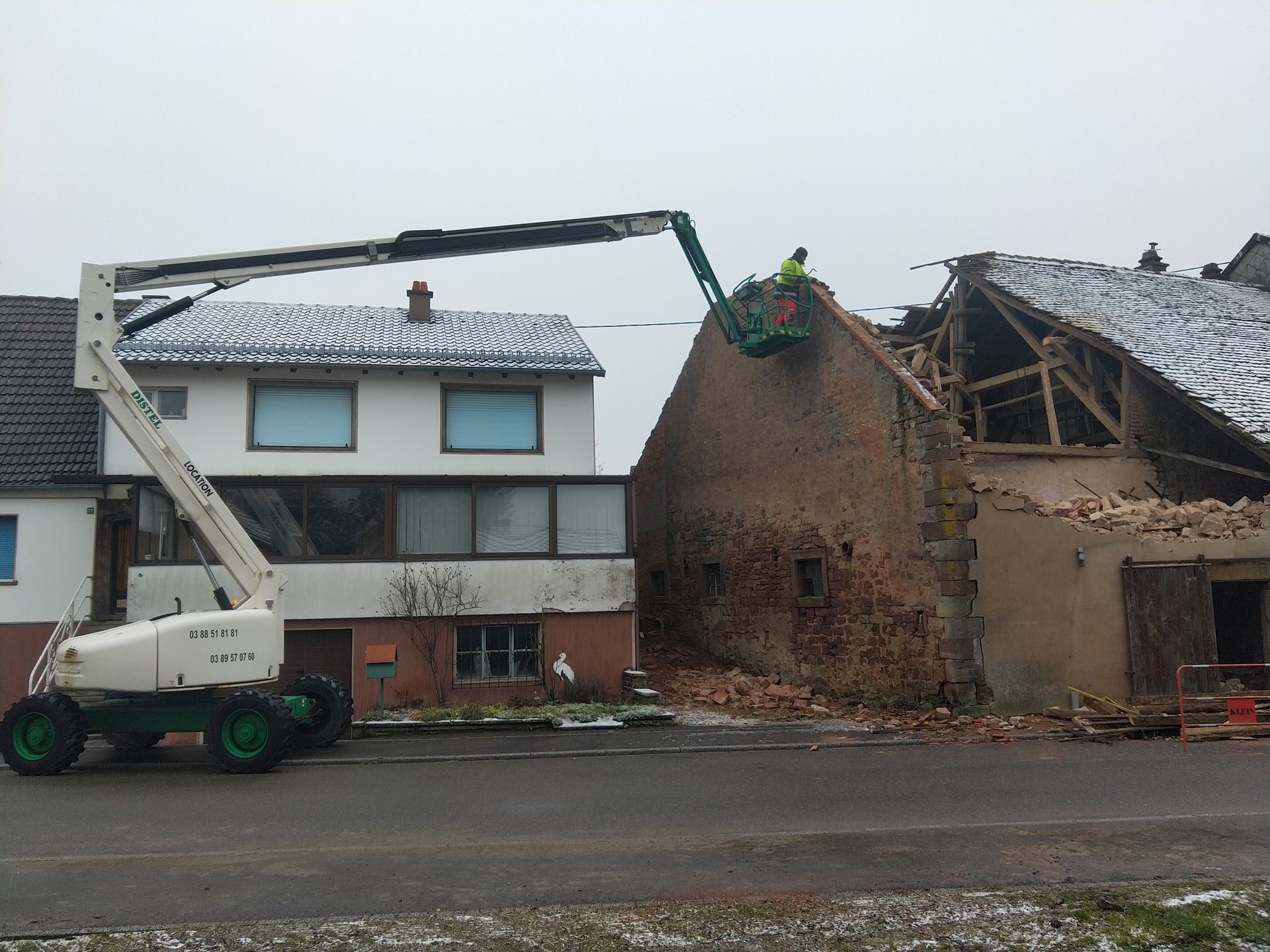 Une grue démolit une maison à côté d'un bâtiment en briques endommagé.