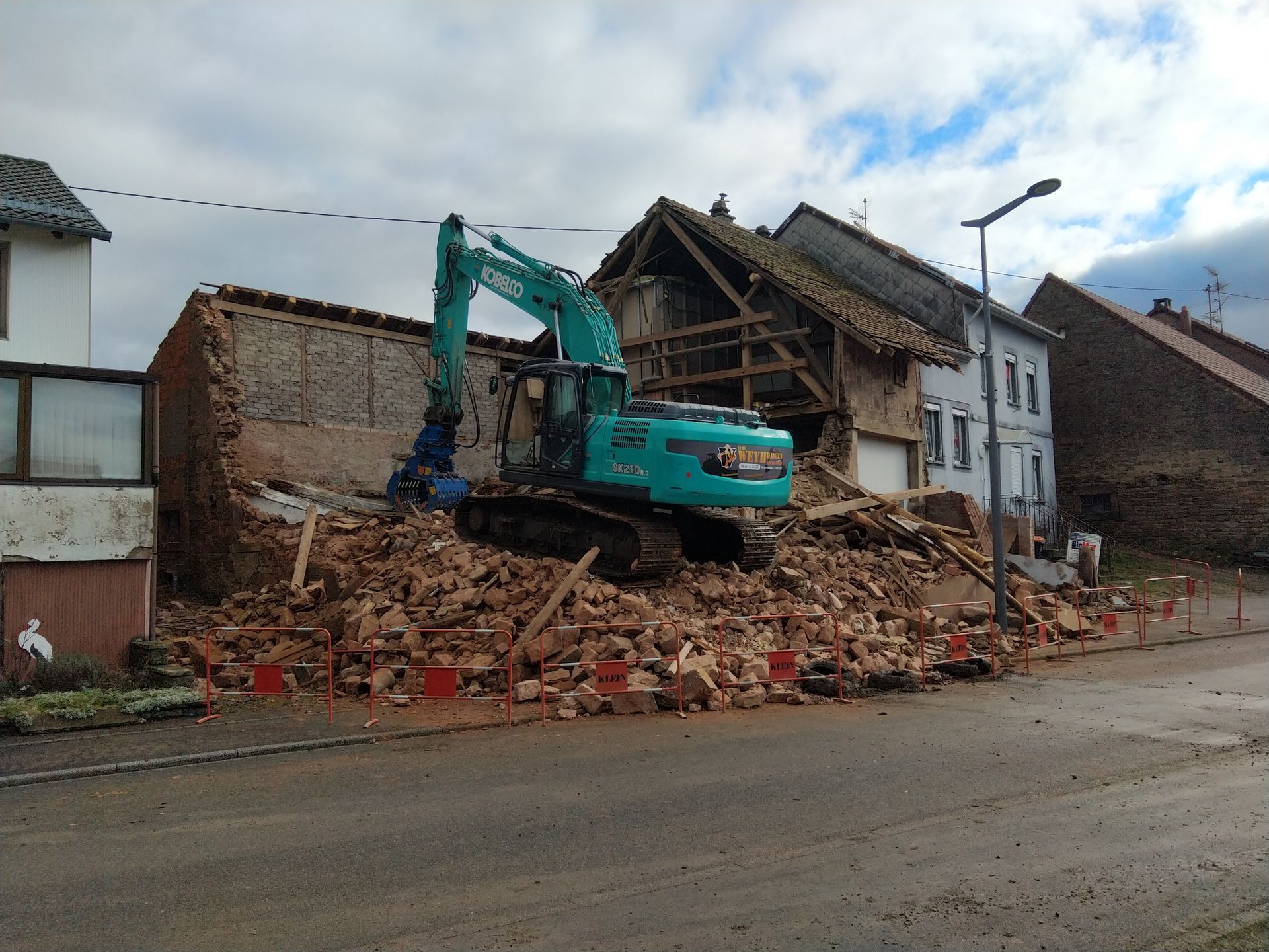 Une pelleteuse turquoise démolit une maison en briques en bordure de rue, des gravats s'entassent devant.