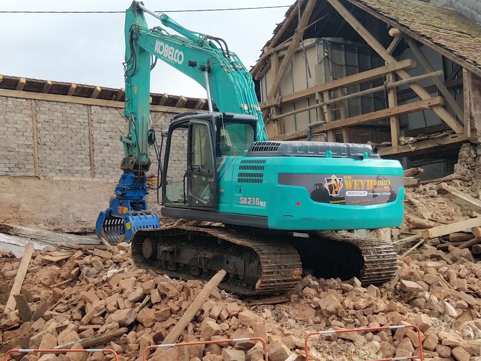Chantier de démolition de maisons avec une excavatrice et des gravats entre deux habitations.