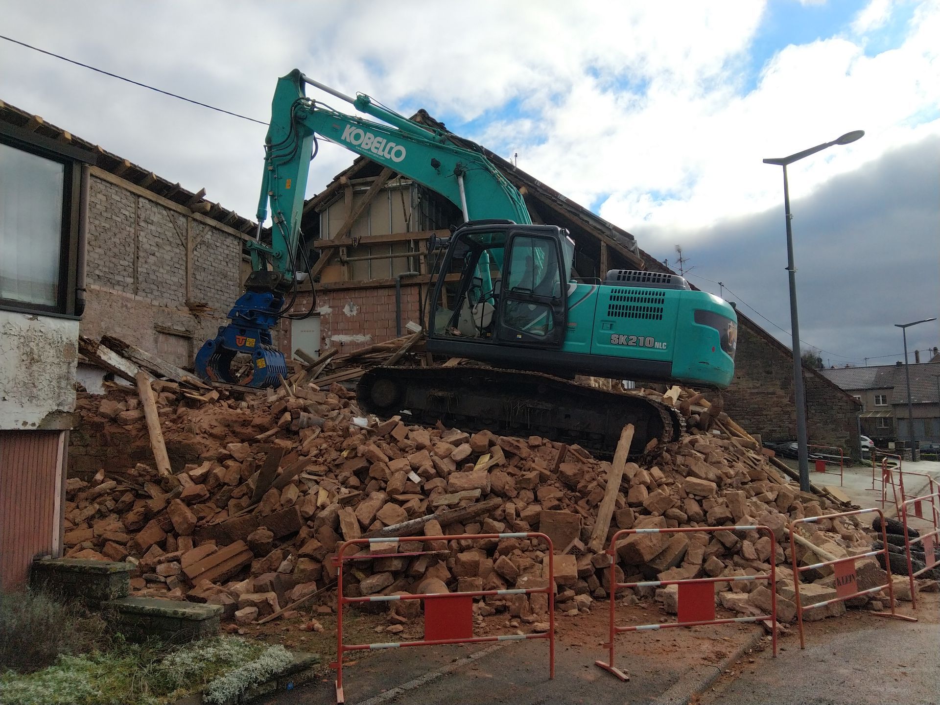Une pelleteuse turquoise sur des décombres à côté d'un bâtiment en briques effondré sous un ciel nuageux.