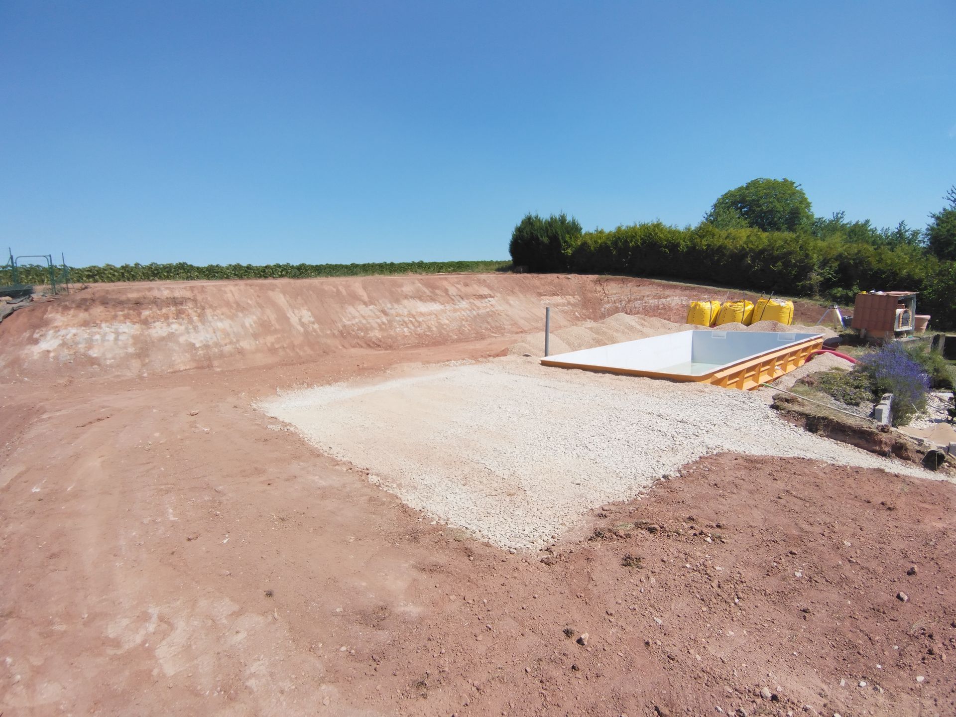 Chantier de construction en gravier avec dalle de béton, remblai de terre et arbres sous un ciel bleu clair.