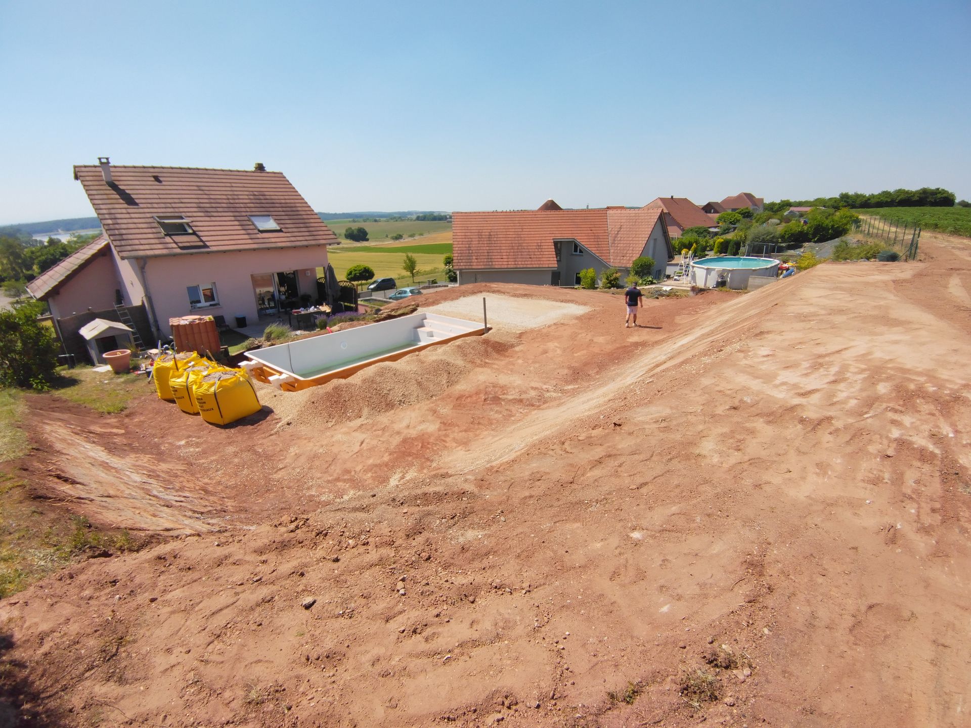 Chantier de construction rural avec de nouvelles maisons, un chemin de terre et des engins de chantier jaunes sous un ciel dégagé.