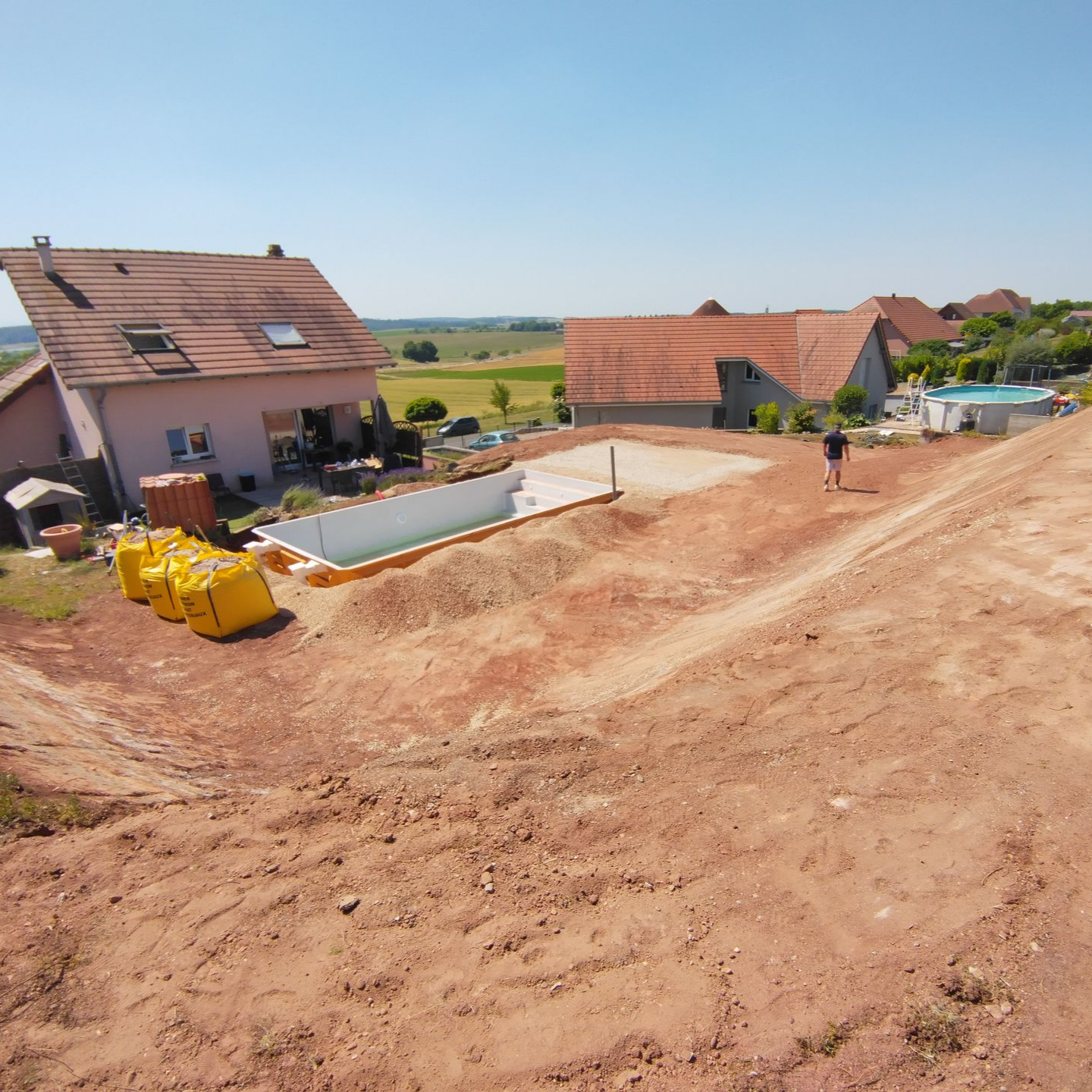Chantier de construction avec maisons neuves, terrain vague, dalle de béton et ouvriers par une journée ensoleillée.