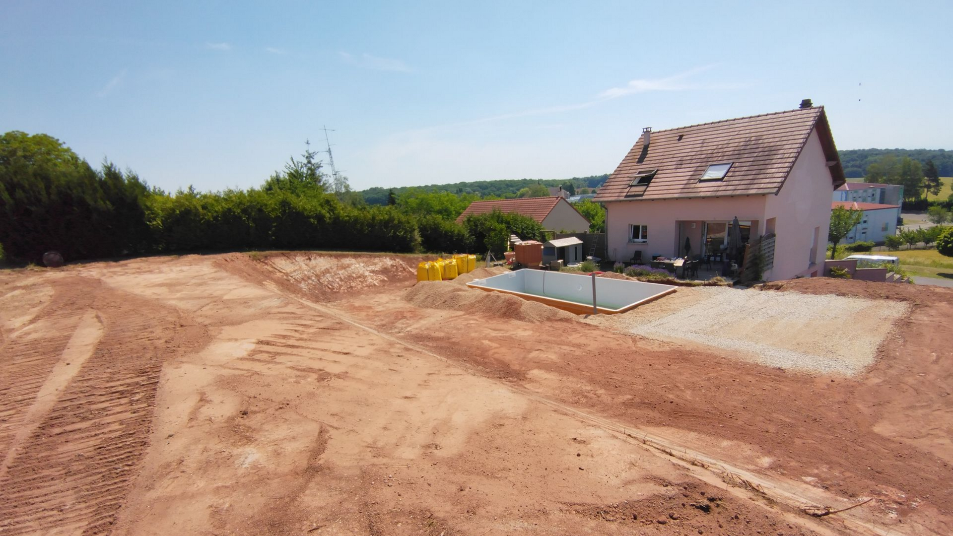 Vaste chantier de terre battue à côté d'une maison au toit rouge sous un ciel bleu azur.