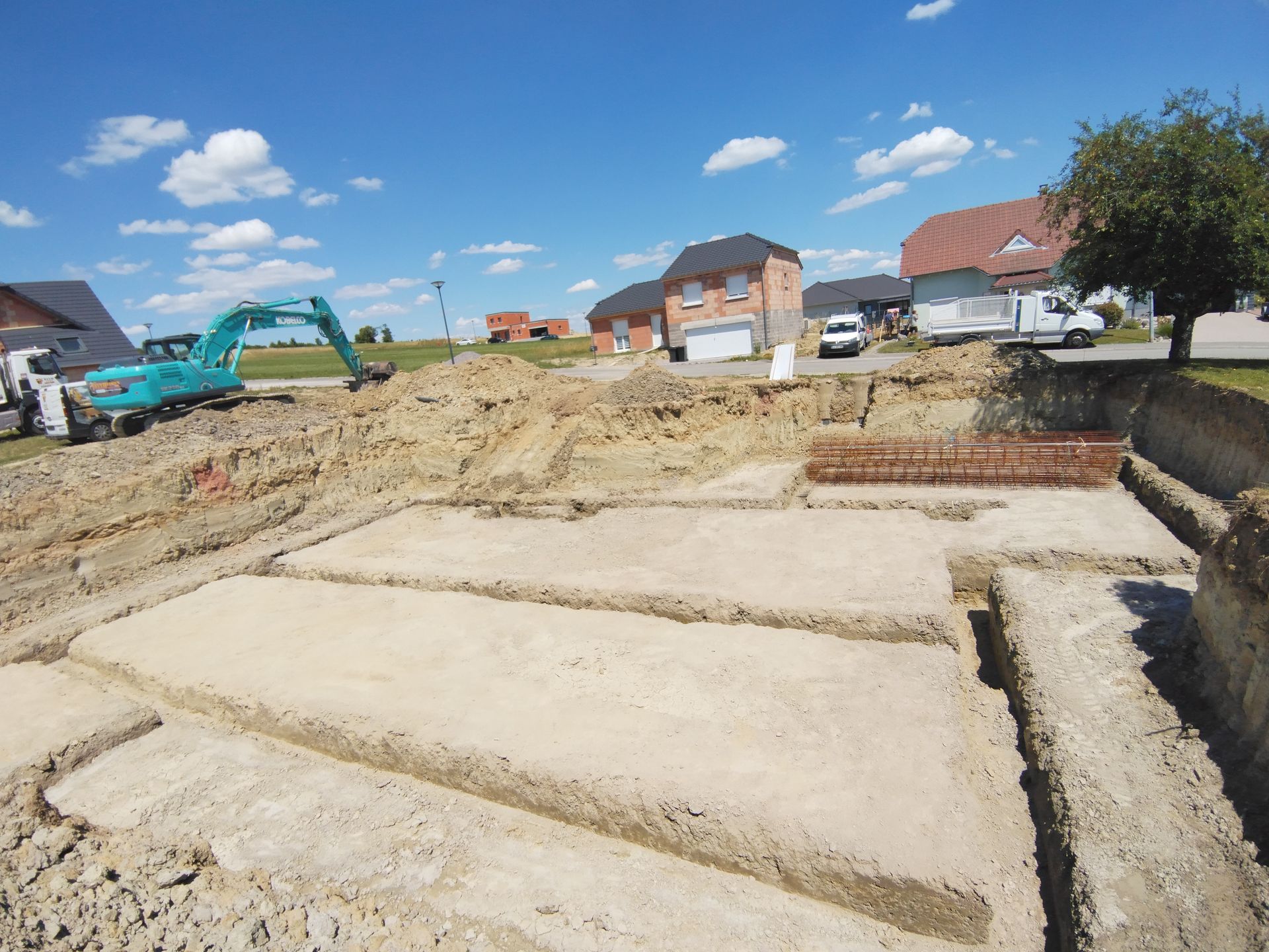 De nouvelles maisons sont en construction, avec des tranchées de fondation en béton et une excavatrice, sous un ciel bleu.