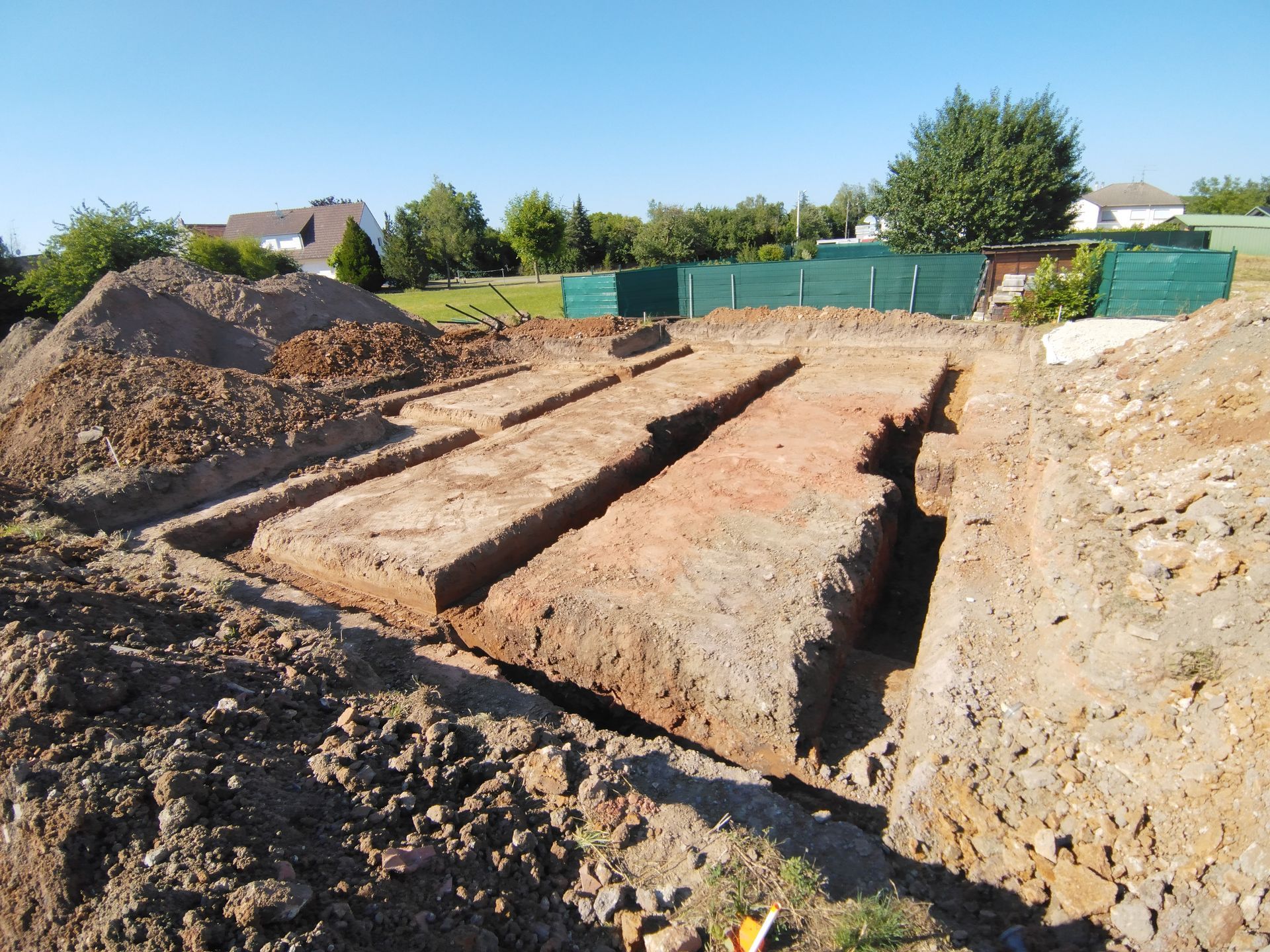 Chantier de fouilles avec coffrages de fondation de tranchées et tas de terre sous un ciel bleu clair.