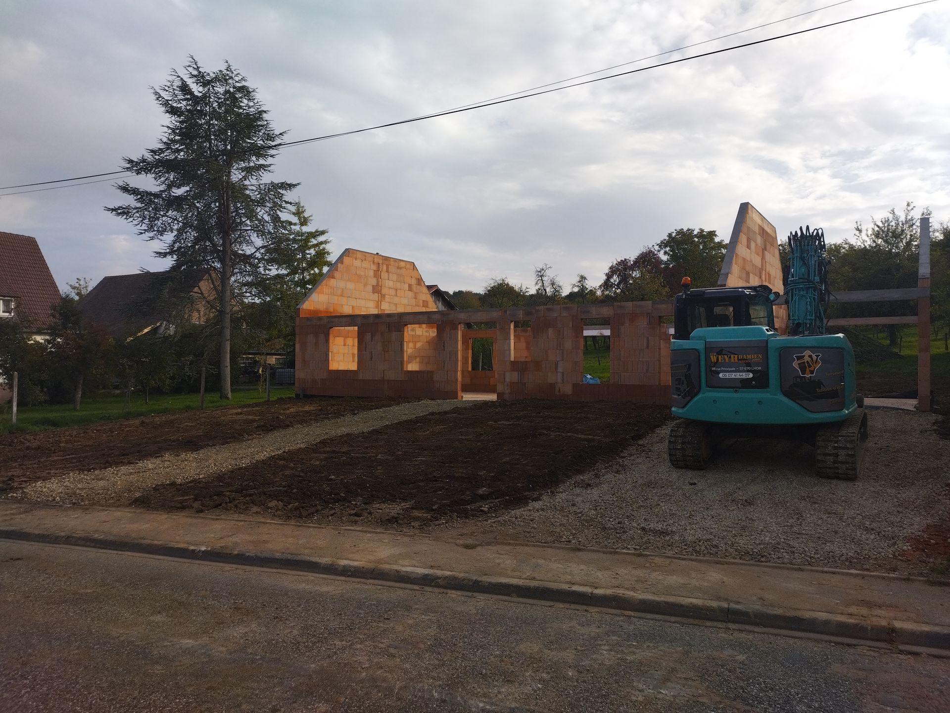 Maison partiellement construite avec une ossature en bois à côté d'une maison blanche terminée sur un chantier boueux.