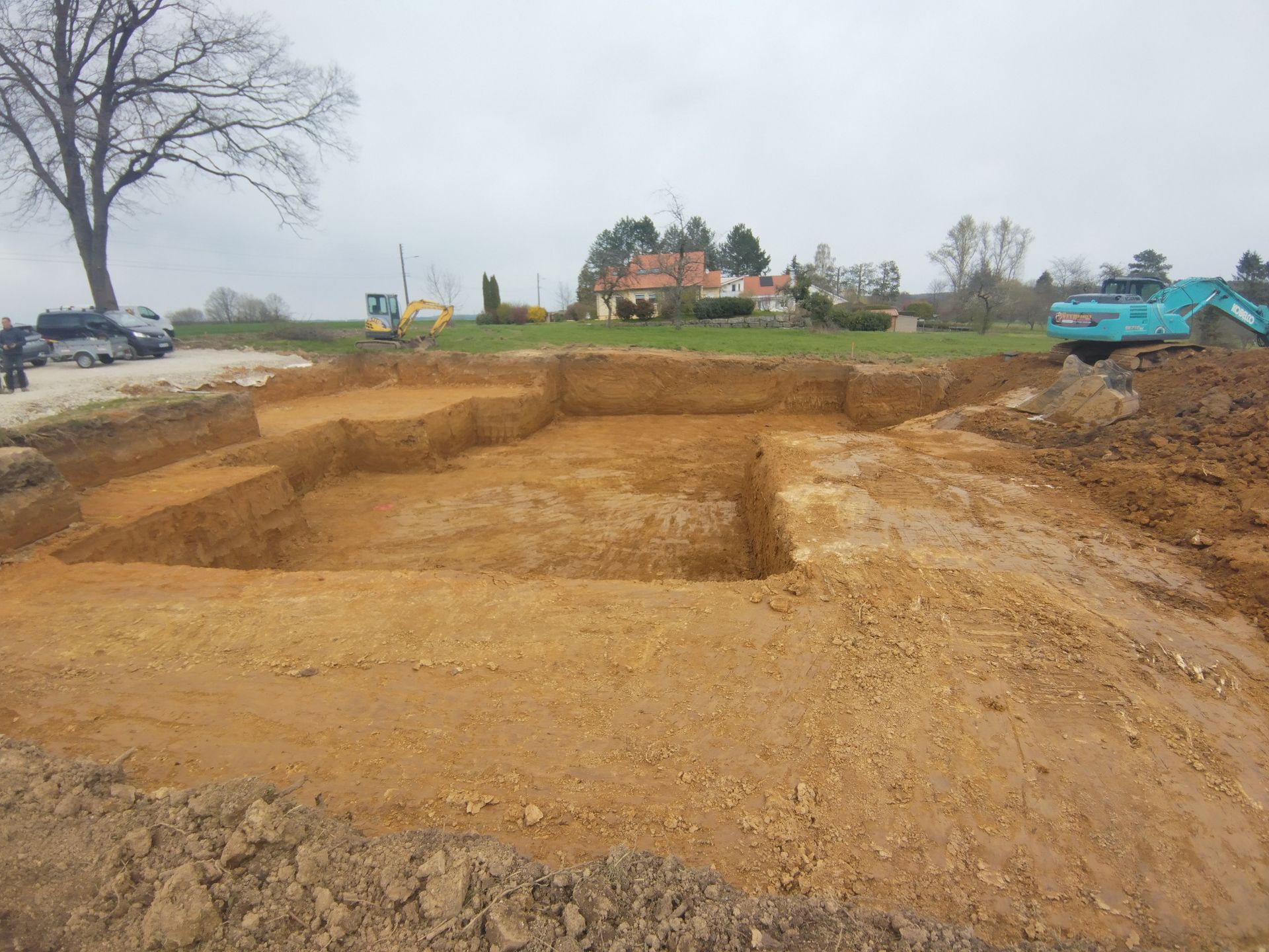 Terrain en terre excavée avec des tranchées étagées, un sol boueux et des engins de chantier sous un ciel couvert.