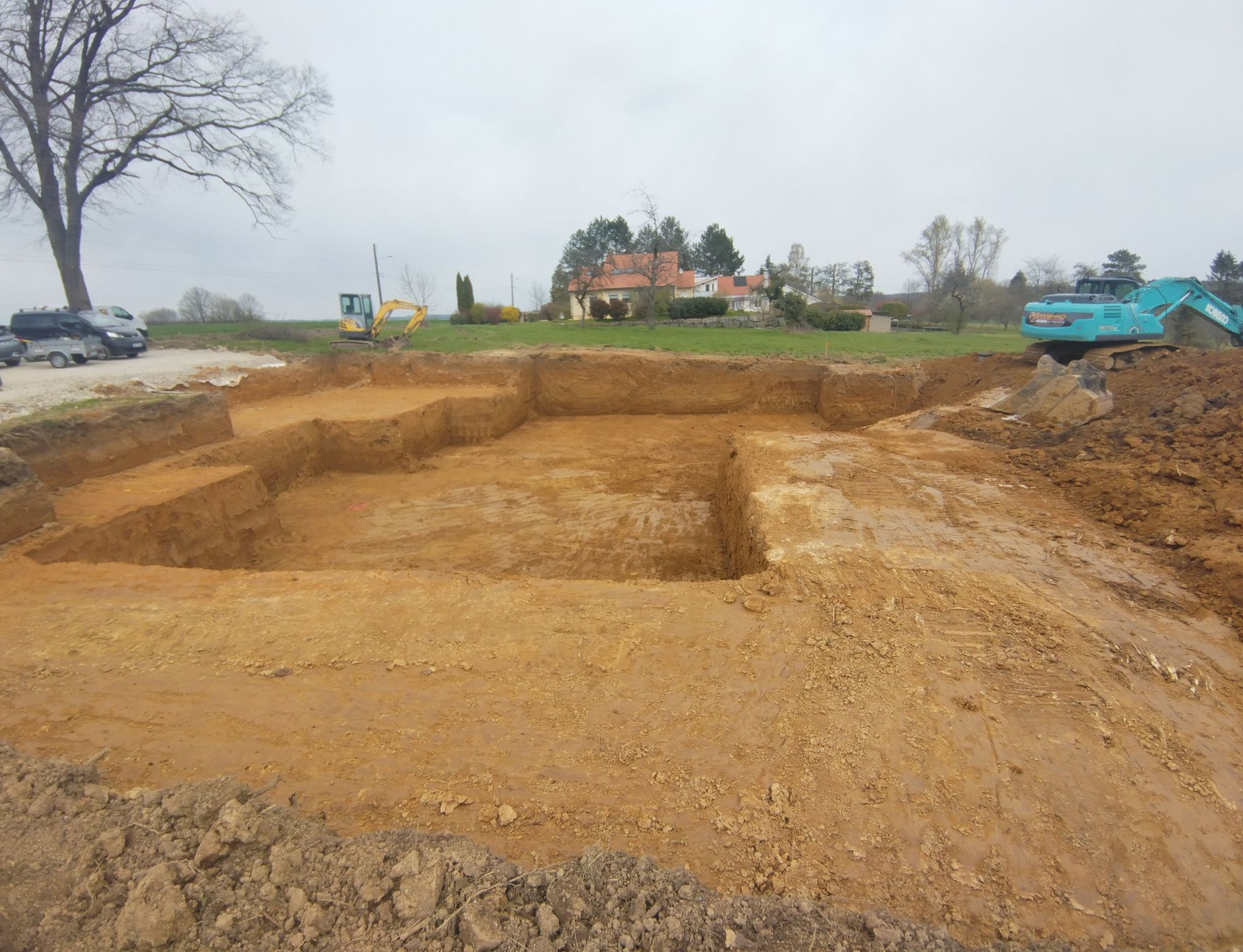Une excavatrice et un bulldozer travaillent sur un chantier boueux, au bord de l'eau et des arbres.
