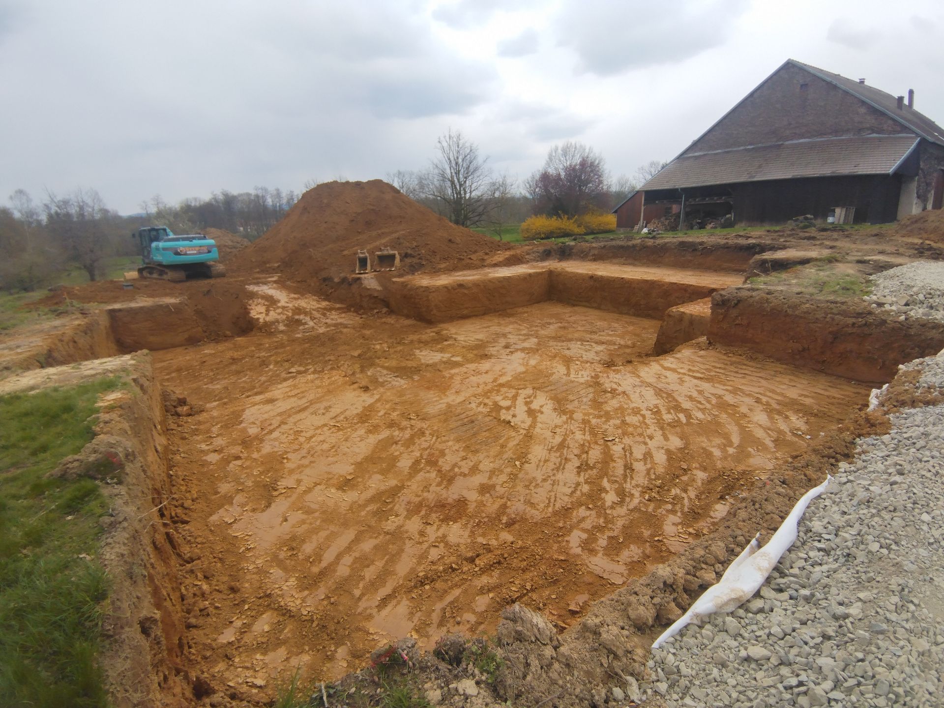Chantier de fondation en terre excavée à côté d'une maison, avec une pelleteuse bleue et des tas de gravier sous un ciel nuageux.
