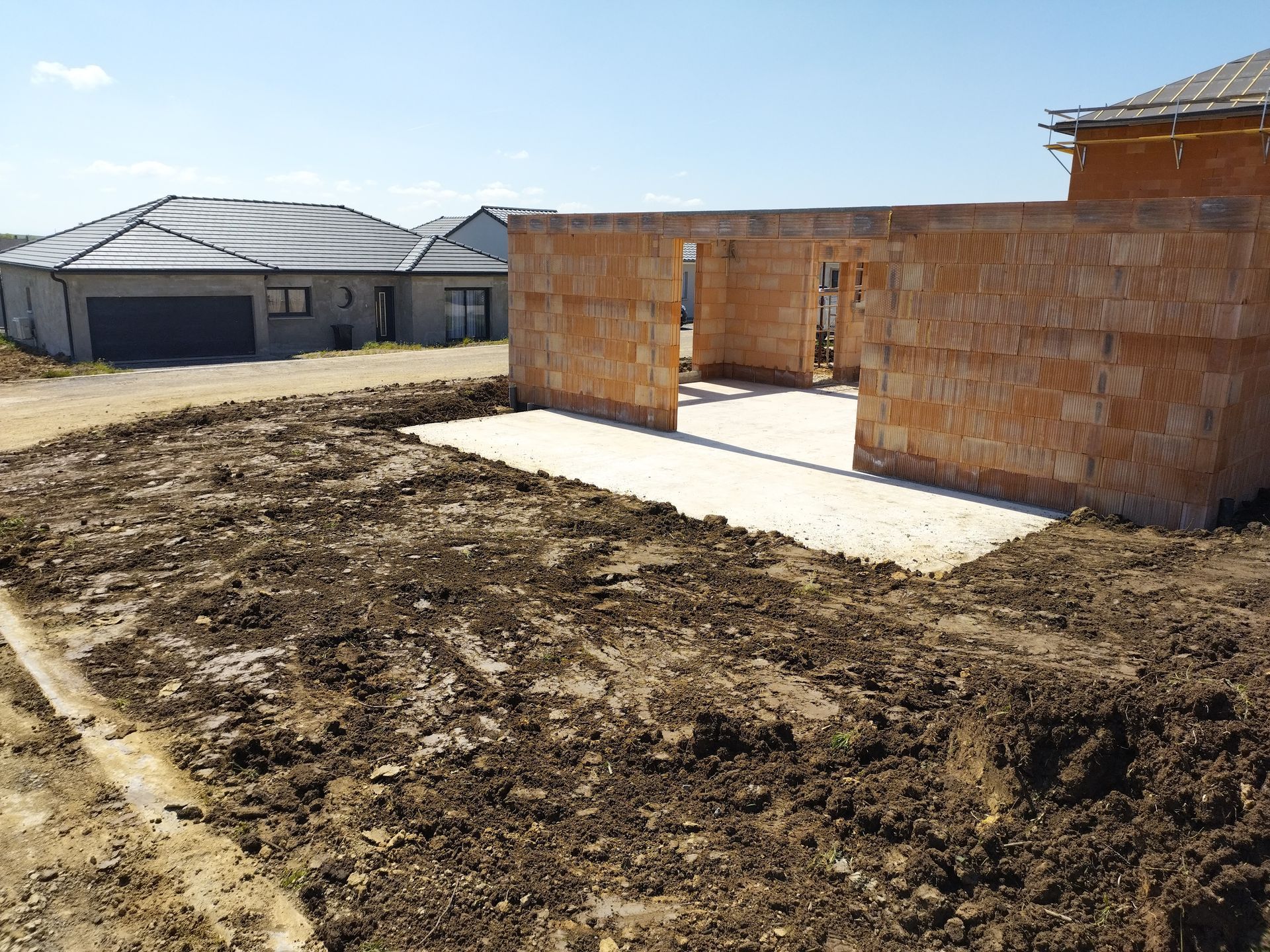 Maison en briques partiellement construite sur un terrain vague, à côté de maisons achevées, sous un ciel bleu azur.