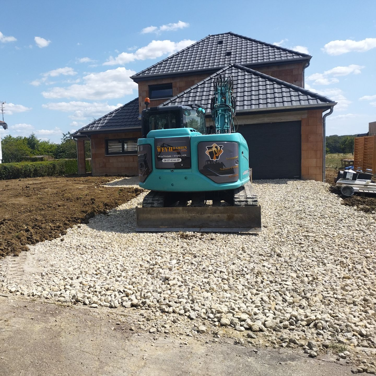 Une pelleteuse turquoise est garée sur une allée de gravier devant une maison en briques en construction.