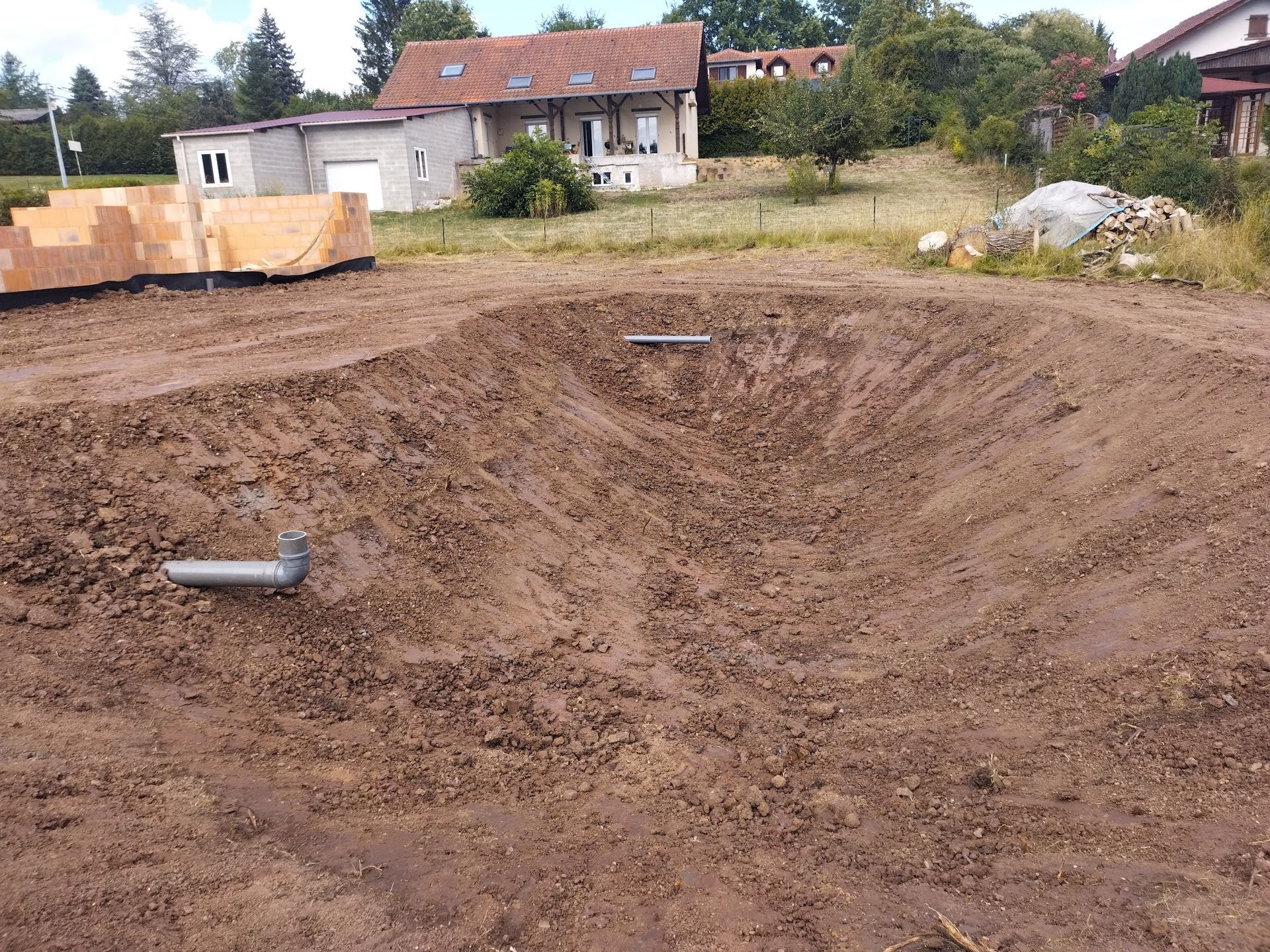 Une excavatrice creuse une tranchée près d'une maison en briques, dans un sol boueux.