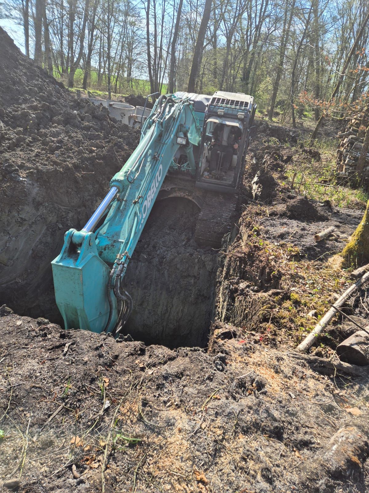Une pelleteuse turquoise creuse une profonde tranchée dans une zone boisée et boueuse, près d'un monticule de terre.