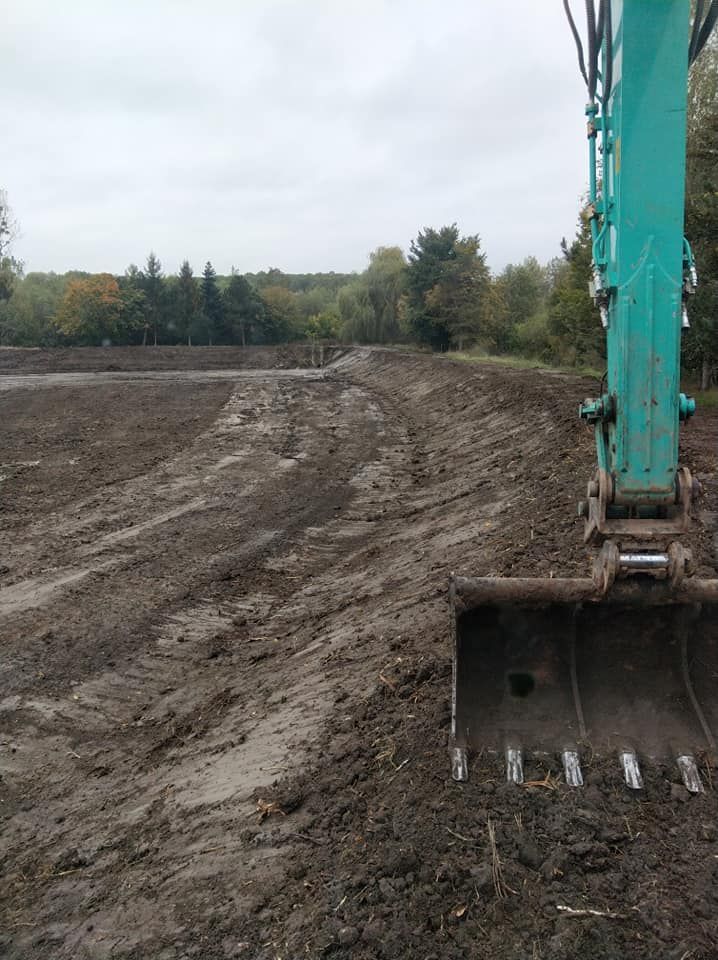 Godet d'excavatrice sur un chantier boueux, près d'une rangée d'arbres, sous un ciel couvert.