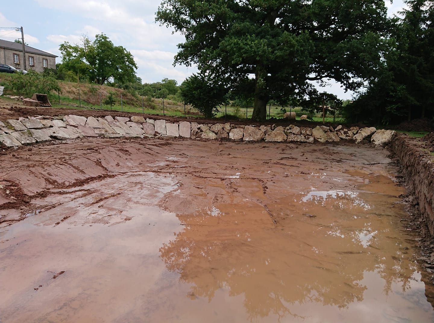 Fosse d'excavation boueuse avec peu d'eau, mur de soutènement en pierre et arbres en arrière-plan.
