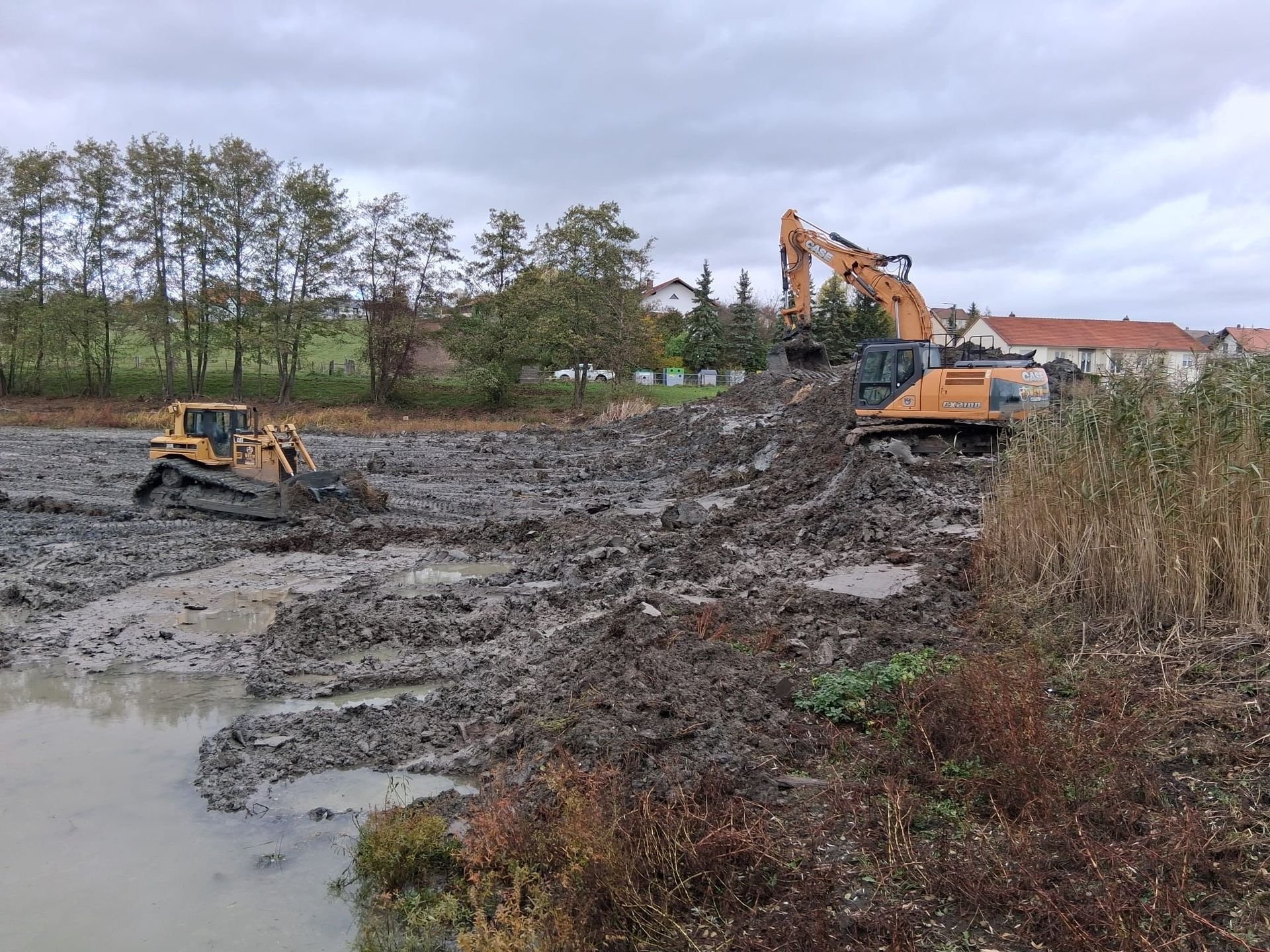 Un bulldozer et une excavatrice déblayent un terrain boueux près d'un étang, avec des arbres et des maisons en arrière-plan.