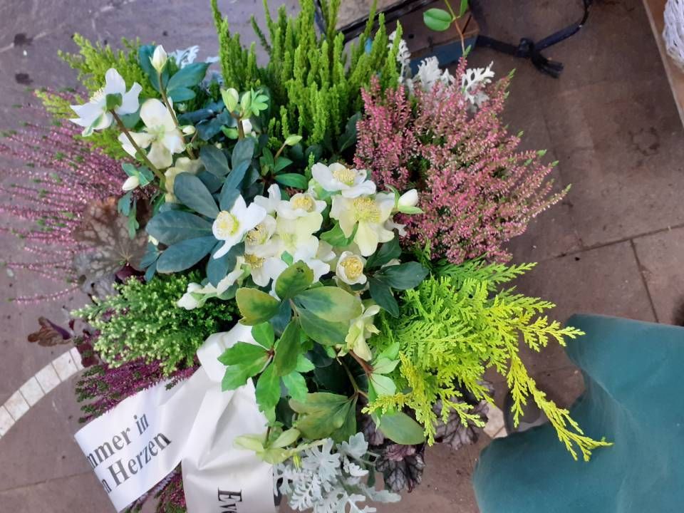 Blumenarrangement mit weißem Band: grüne, rosa und weiße Blumen in einem Korb. Das Band ist beschriftet.