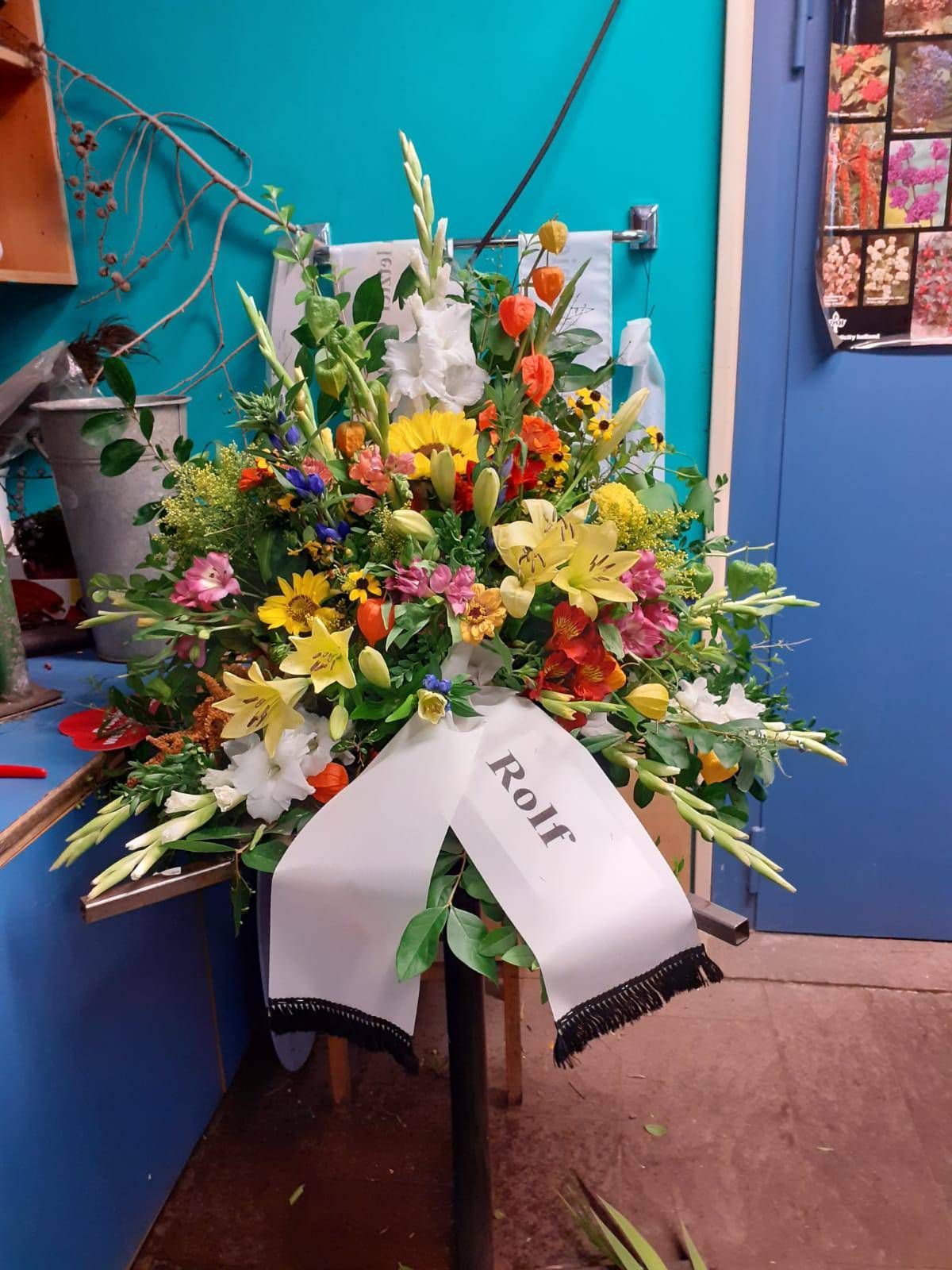 Farbenfrohes Blumenarrangement mit weißem Band und der Aufschrift „Rolf“.