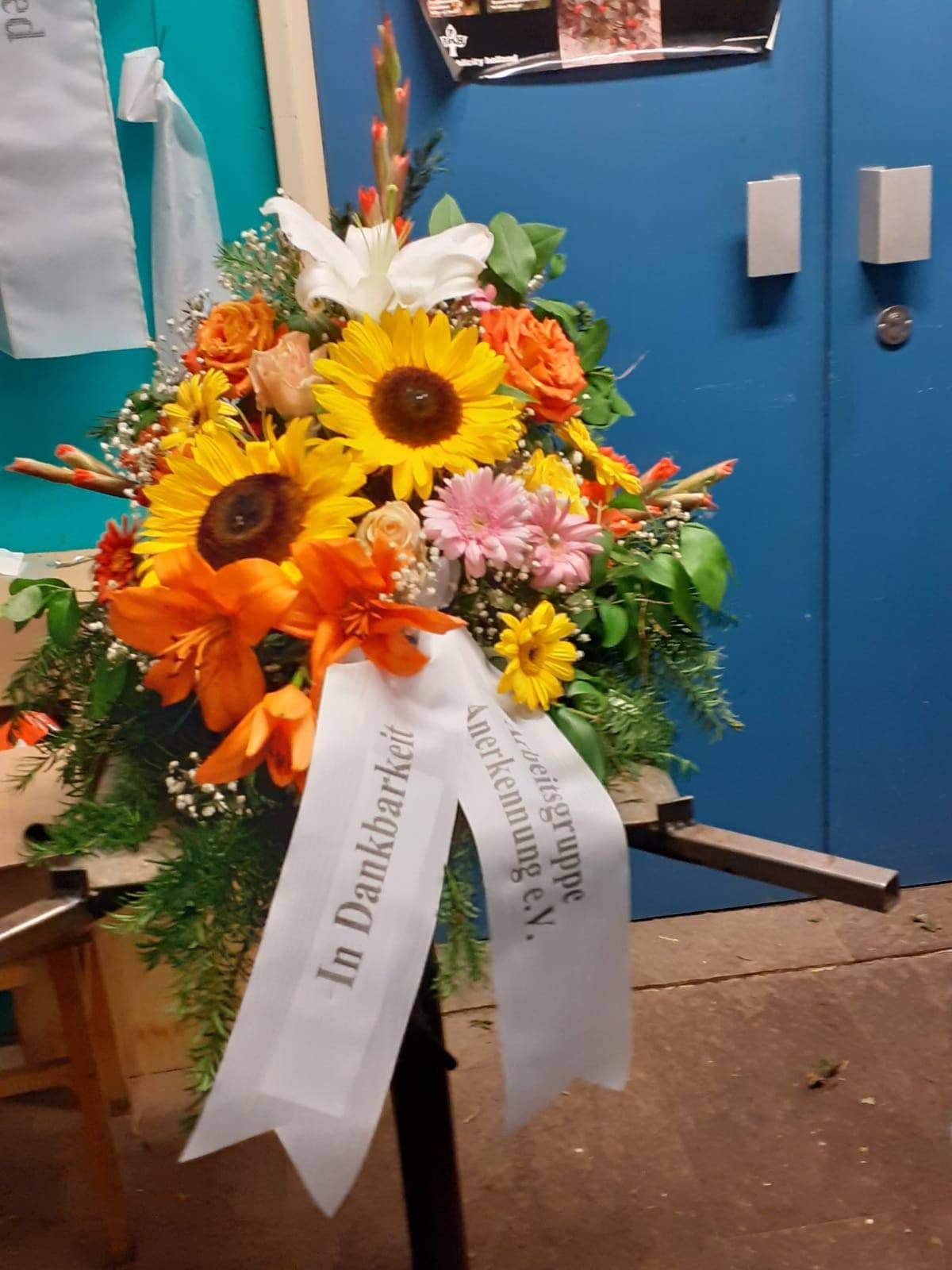 Blumenarrangement mit Sonnenblumen, orangefarbenen Lilien und einem weißen Band mit der Aufschrift „In Dankbarkeit“.