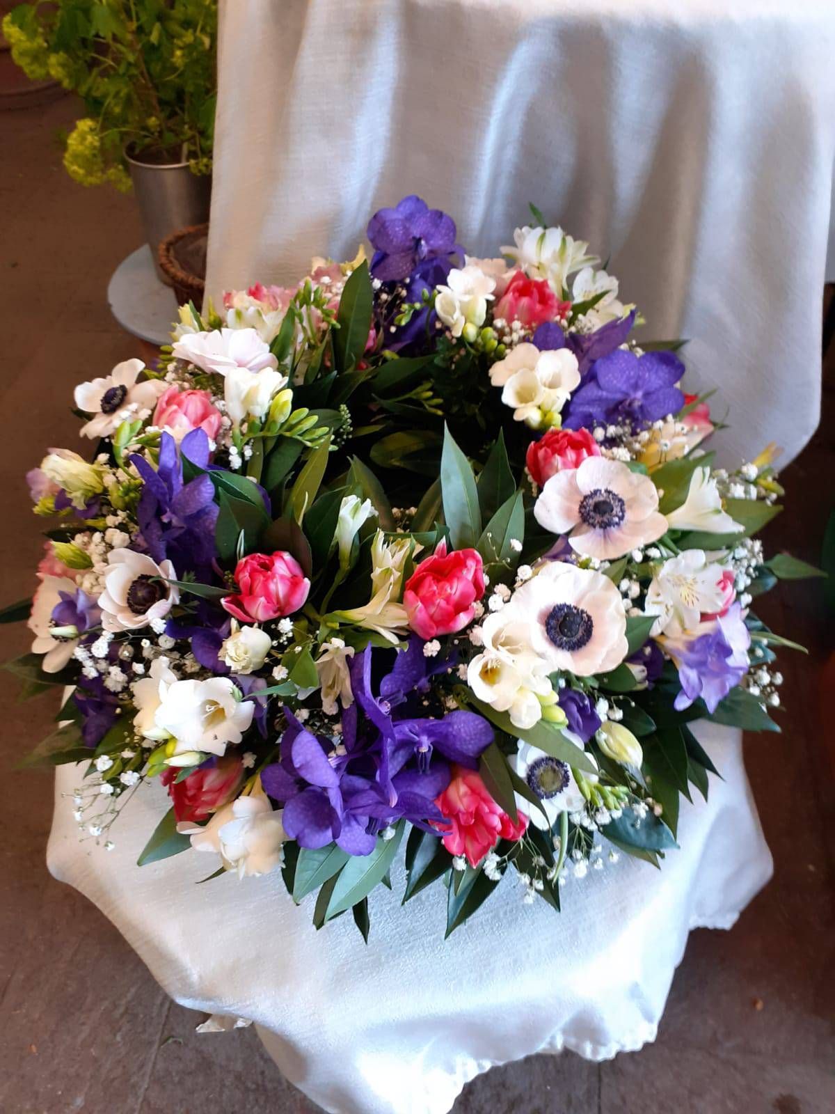 Blumenkranz mit lila, weißen und roten Blüten auf weißem, drapiertem Stoff.