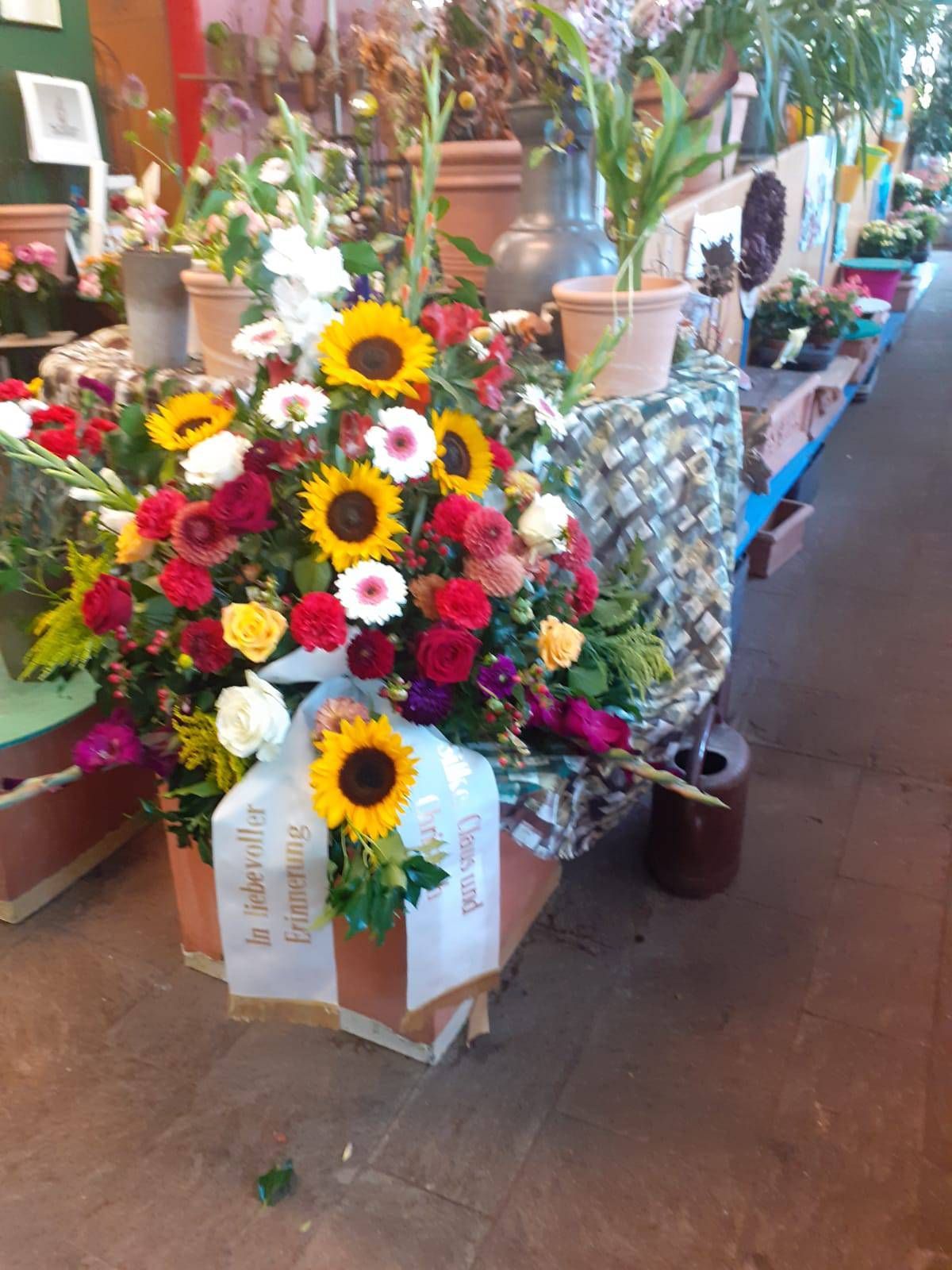 Farbenfrohes Blumenarrangement mit Sonnenblumen, Rosen und einem Band, wahrscheinlich für eine Gedenkfeier, in einem Blumenladen.