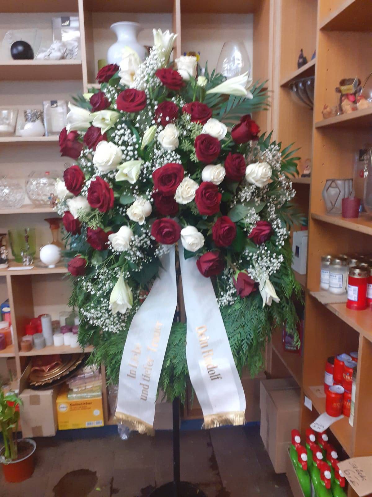 Trauerkranz mit roten und weißen Rosen, weißem Band, in einem Geschäft aufgestellt.