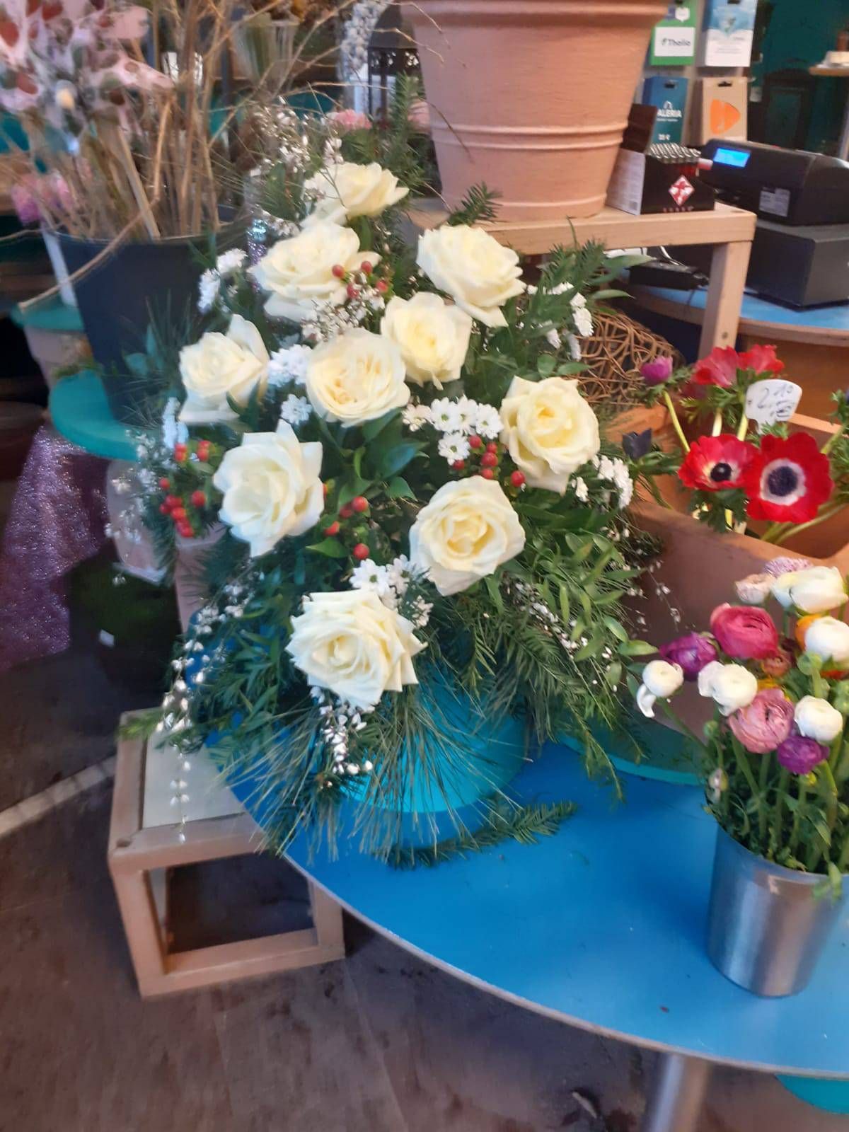Weiße Rosen in einer blauen Vase, umgeben von Grünzeug, auf einem blauen Tisch in einem Blumenladen.